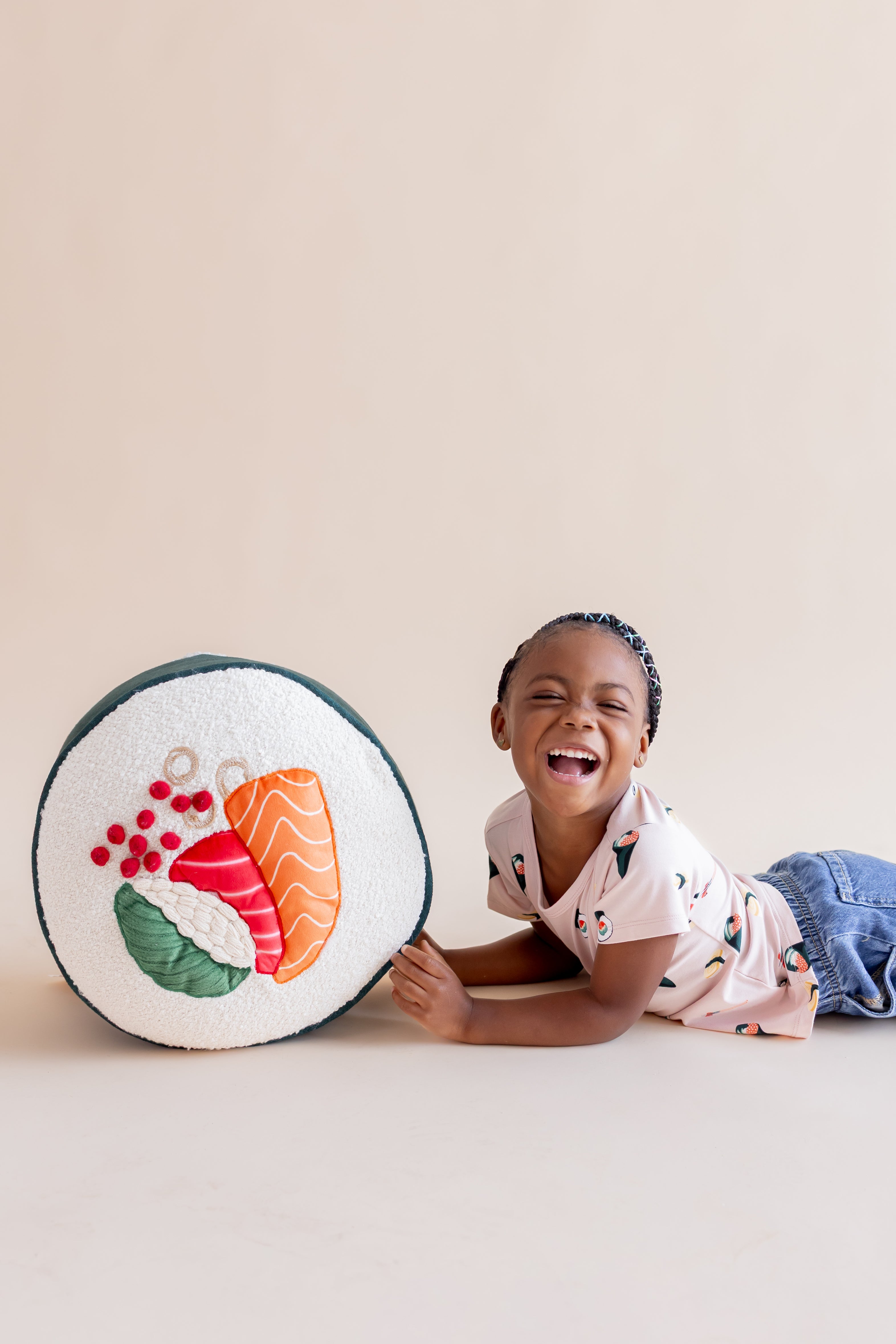 Child laughing on their tummy with blush sushi tee, jeans, and hold a play sushi pillow