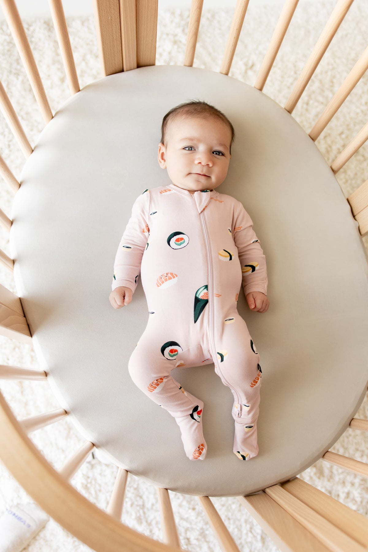 Baby lying in crib wearing Zippered Footie in Blush Sushi print