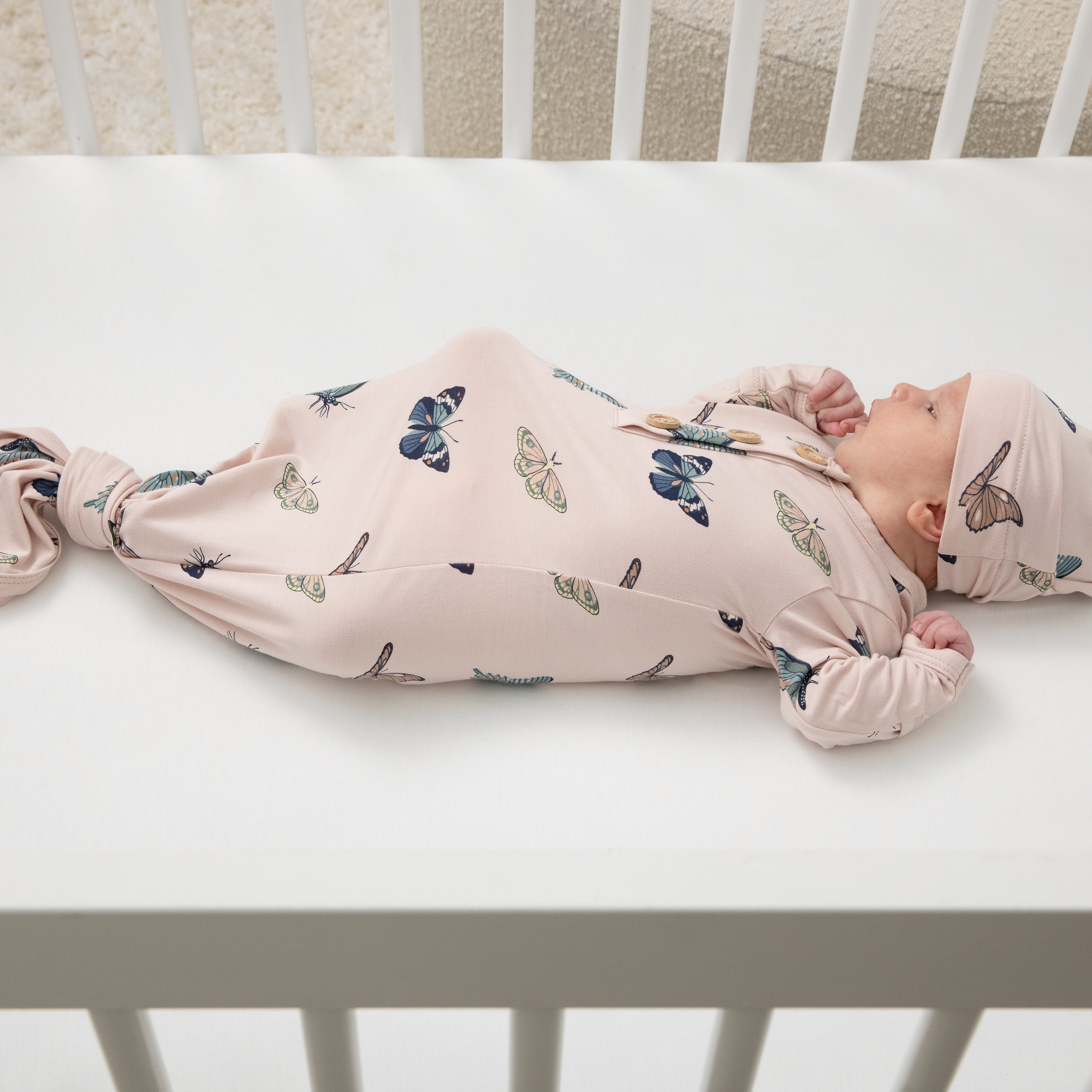 Newborn laying in a crib on a white crib sheet wearing the Knotted Gown with Hat Set in Blush Butterfly