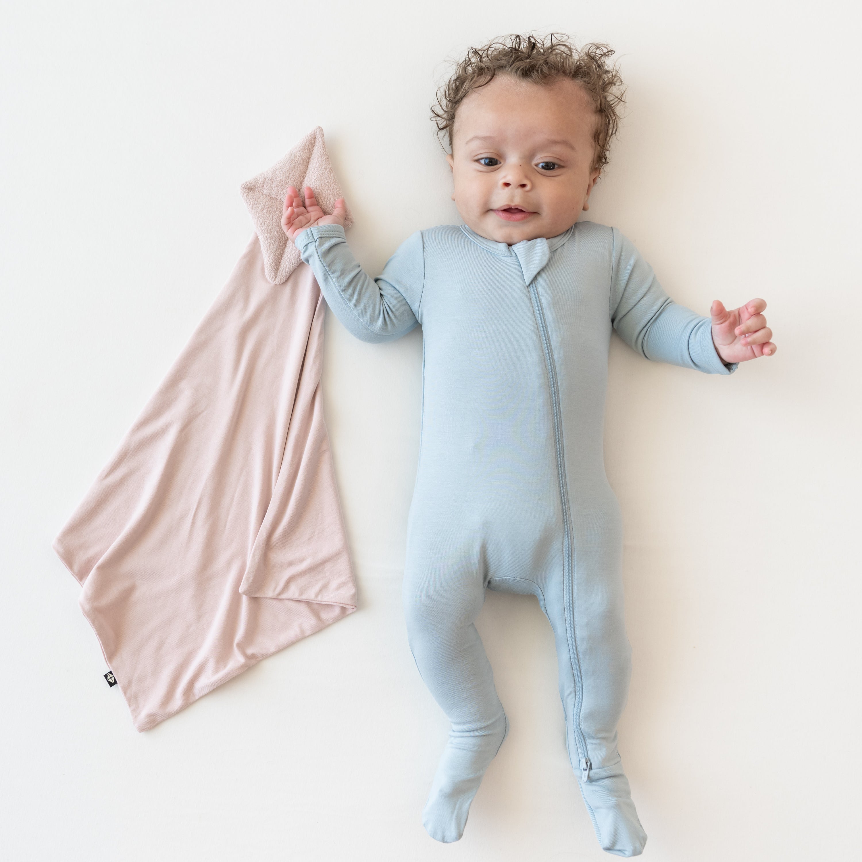 Baby in zippered footie in fog next to a Kite Lovey in Blush