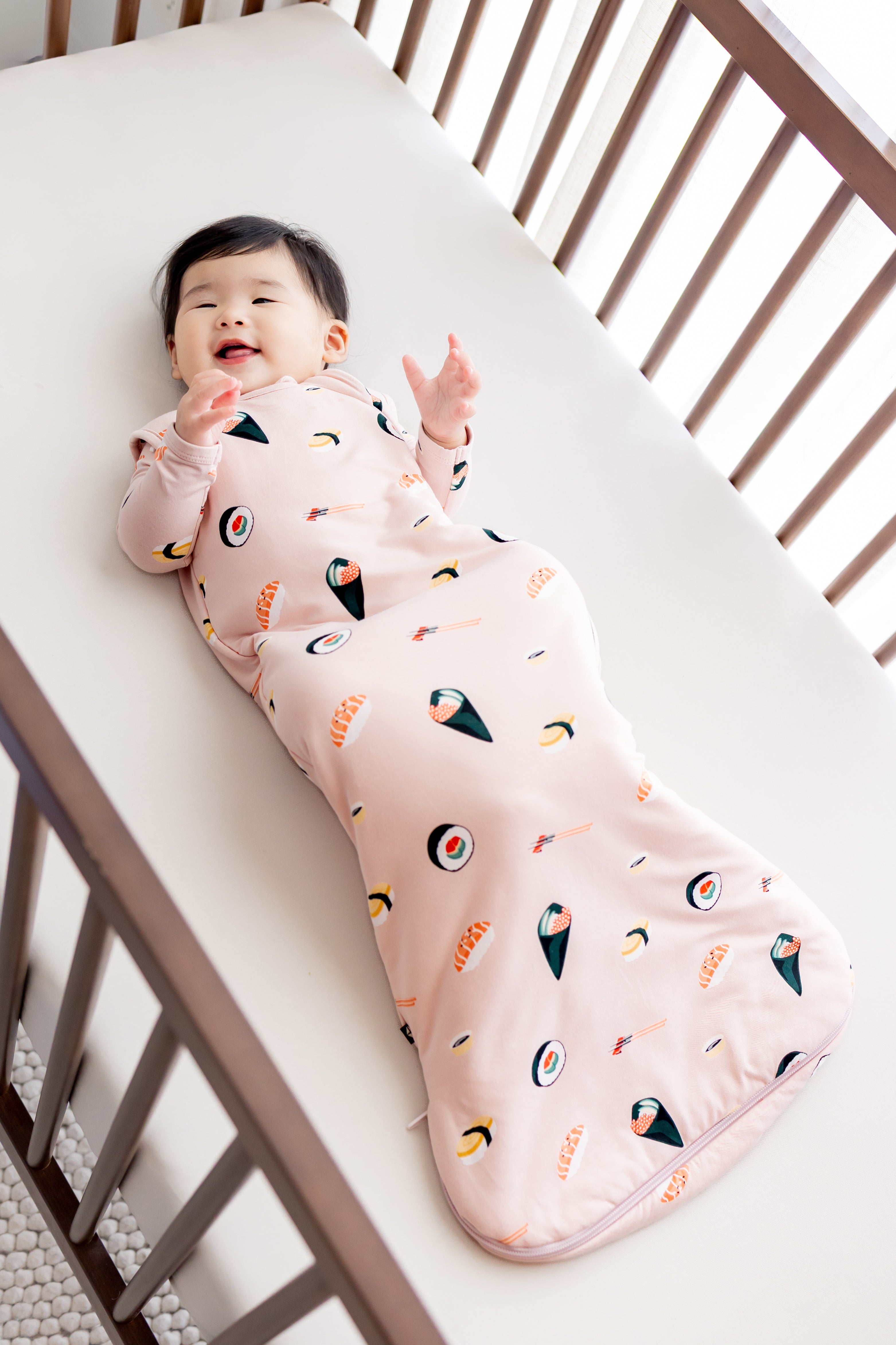Baby laying in crib with white sheets in blush sushi sleep bag