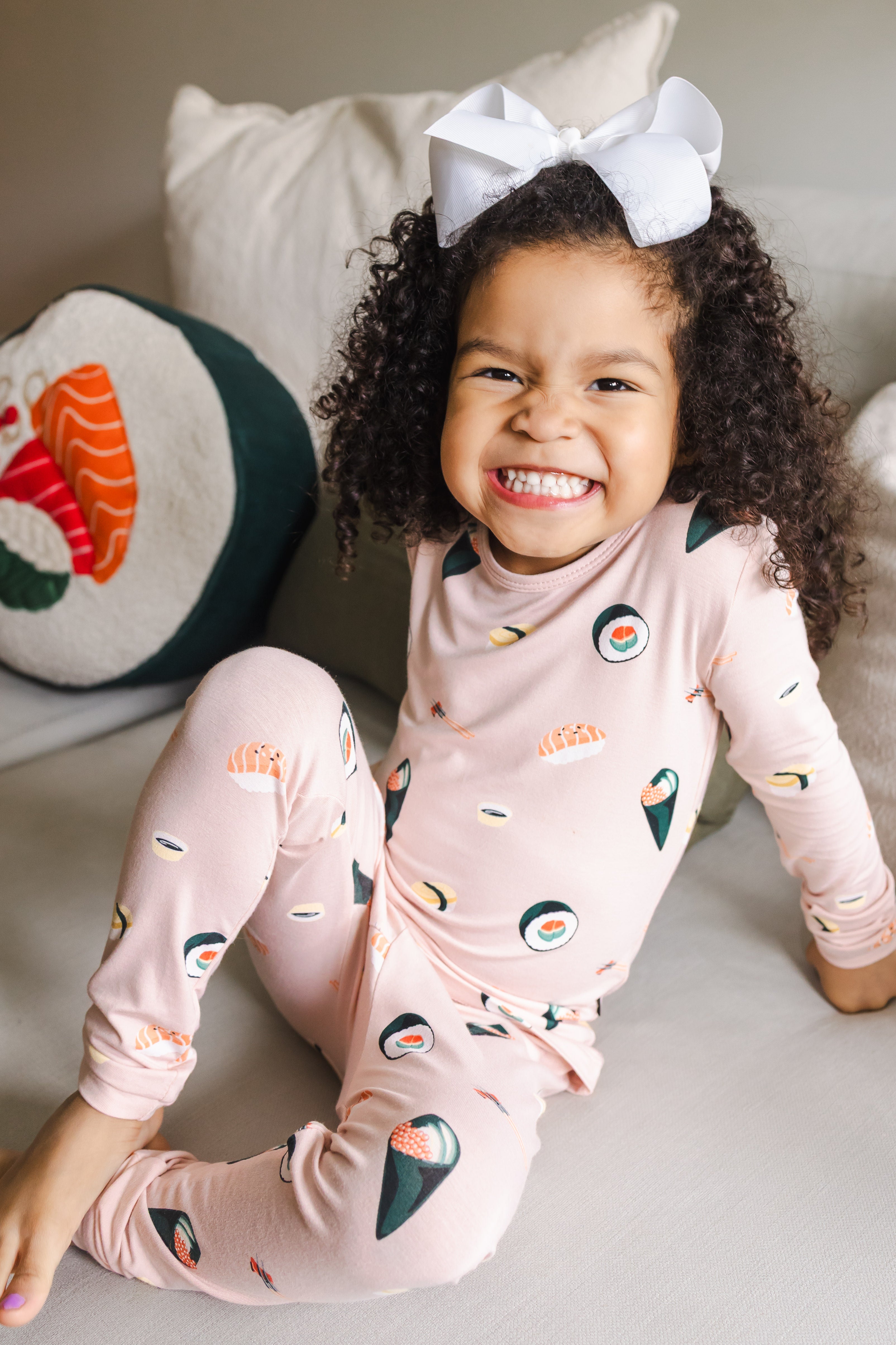 Child smiling sitting on a white couch with one leg up wearing blush sushi pajama set with a white bow
