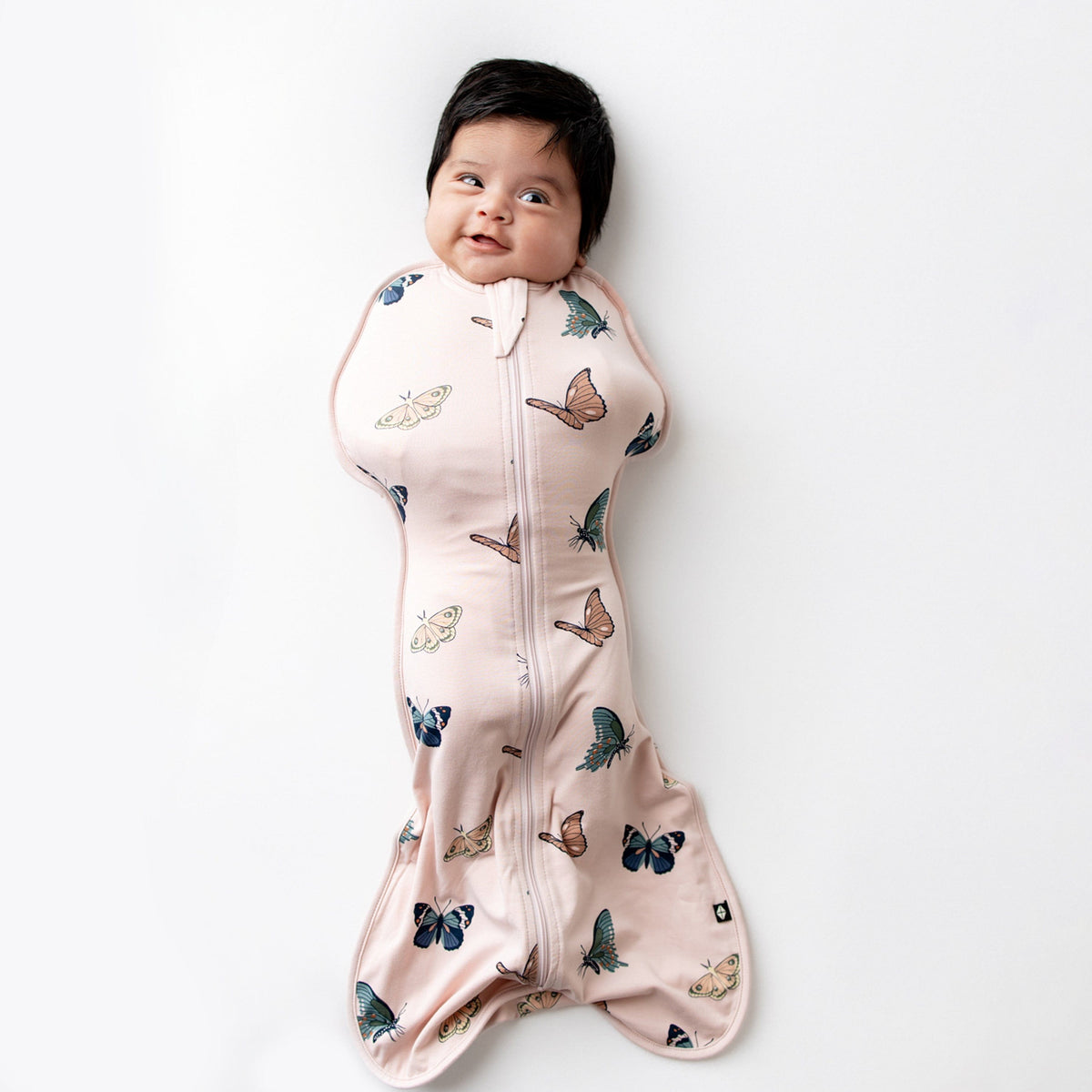 Infant laying on a white hard surface wearing the Swaddle Bag in Blush Butterfly 0.5