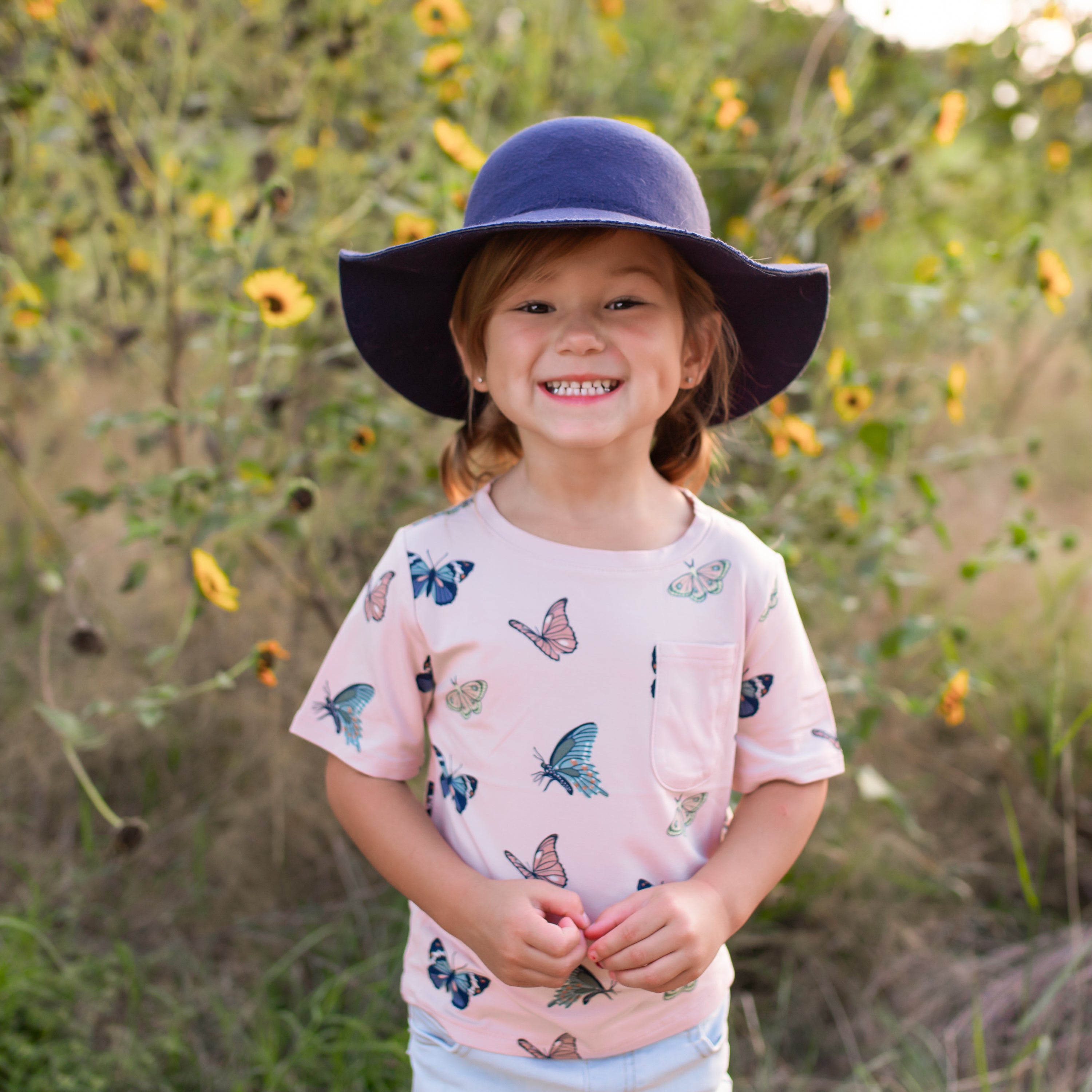 Toddler wearing pink crew neck tshirt with a front pocket in a butterfly print and navy hat outdoors