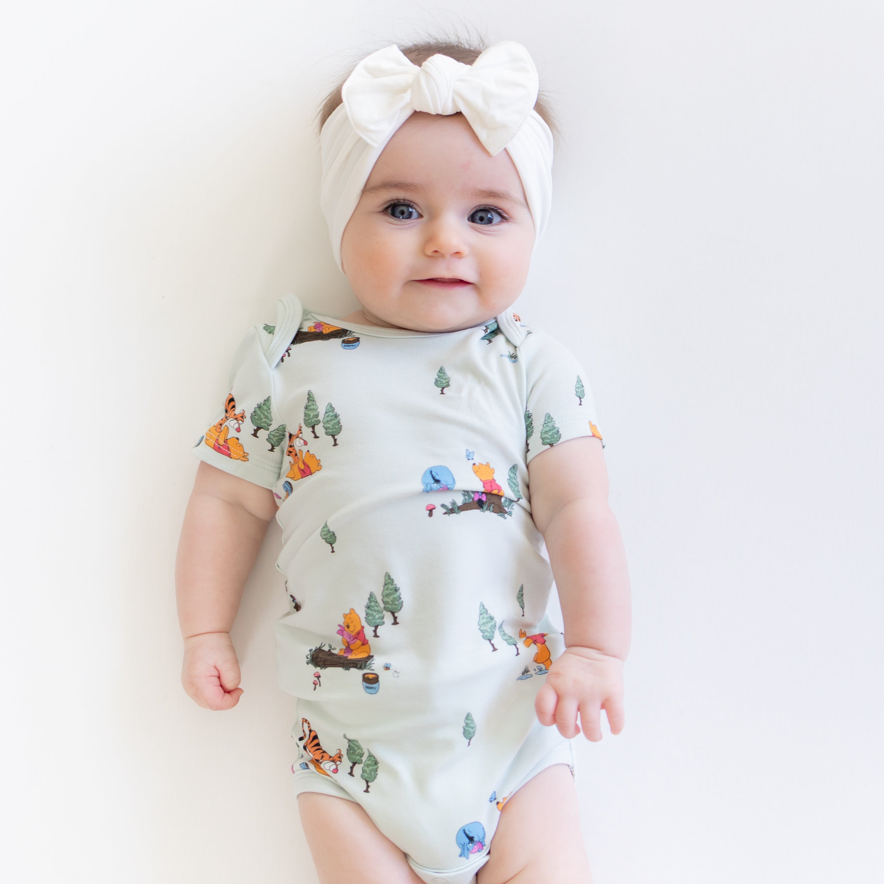 Infant girl wearing the Bodysuit in Winnie the Pooh Hundred Acre Wood paired with a white bow