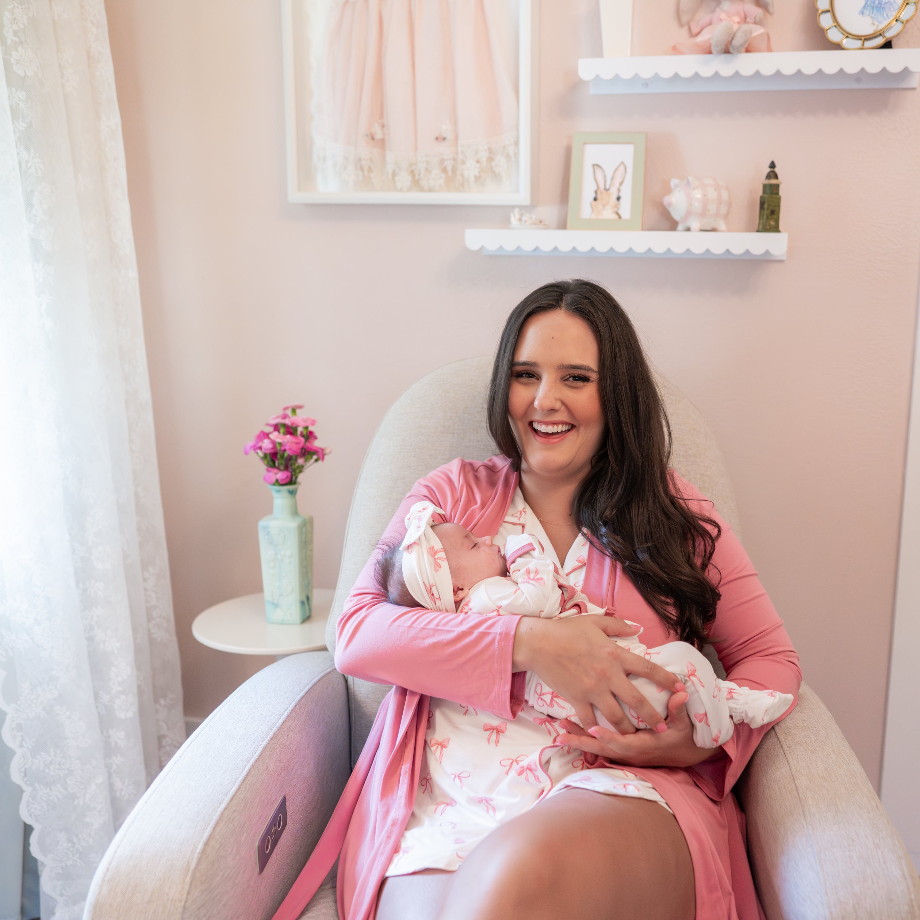Smiling female sitting in a recliner wearing the Women’s Short Sleeve Pajama Set in Bow and Apple Blossom lounge robe holding her newborn matching in the bow print