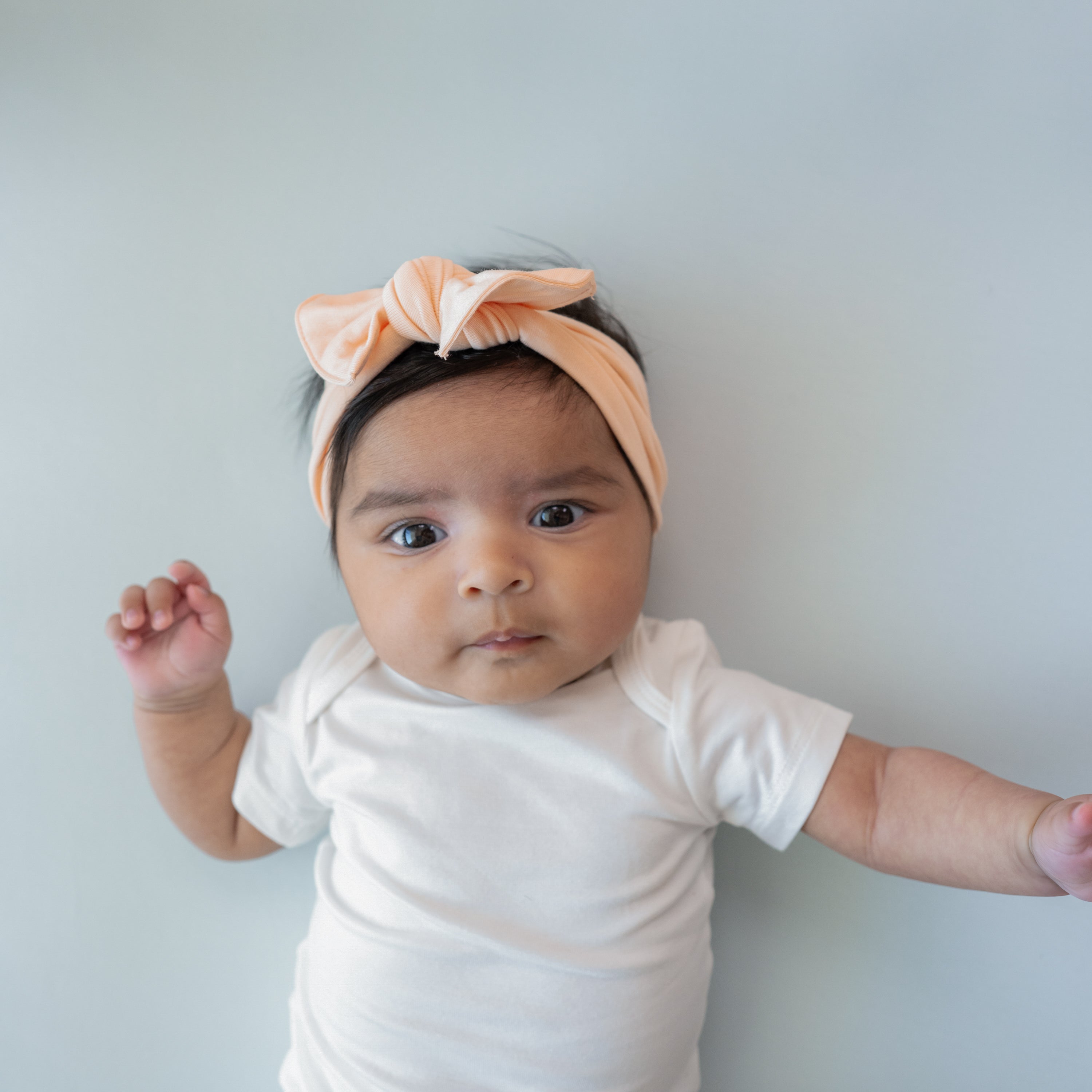 Infant laying down wearing the Knotted Bow Headband in Canyon paired with a Short Sleeve Bodysuit in Cloud