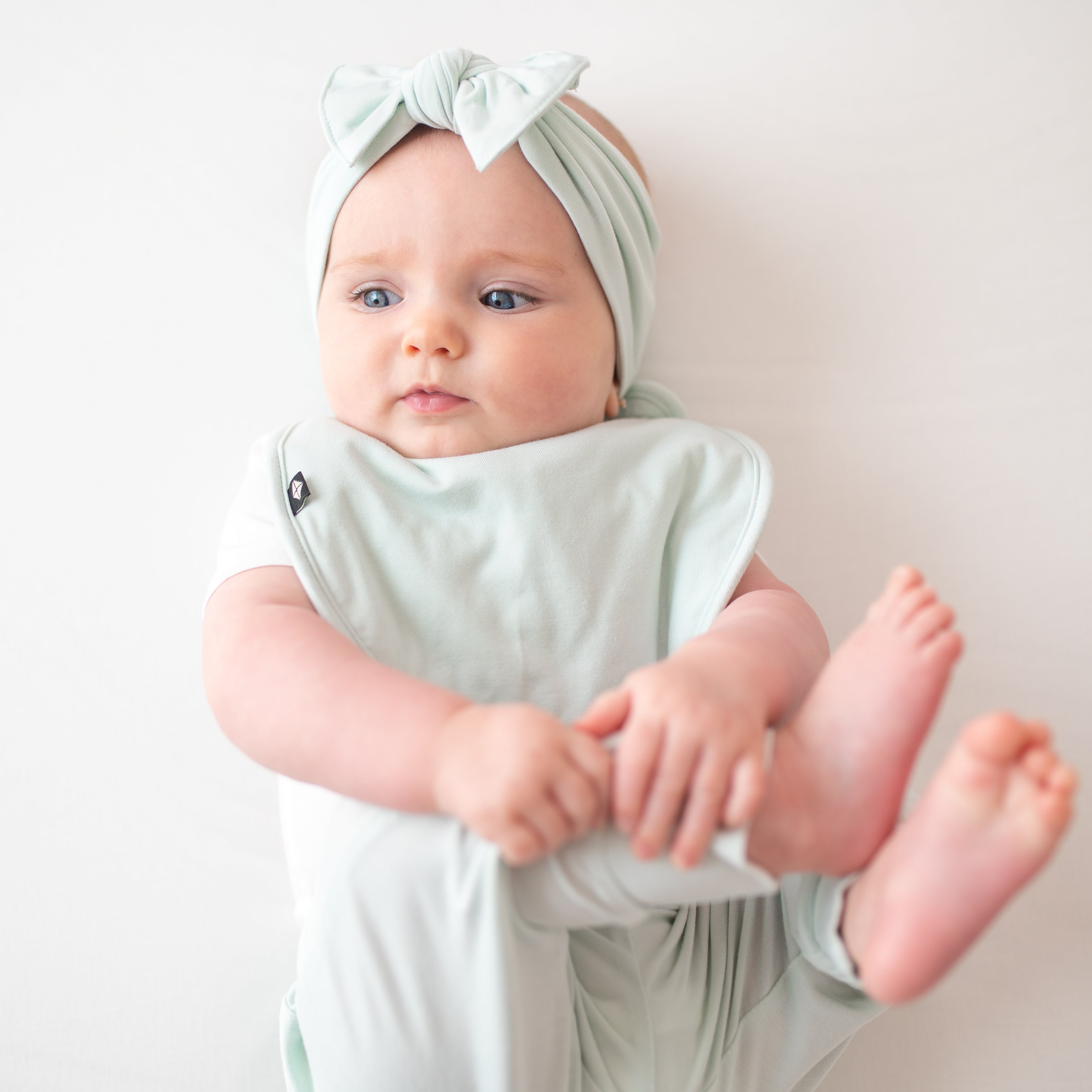 Infant model looking to the side with her feet up in the air wearing the Knotted Bow Headband in Dew with a matching Cushy bib and pants