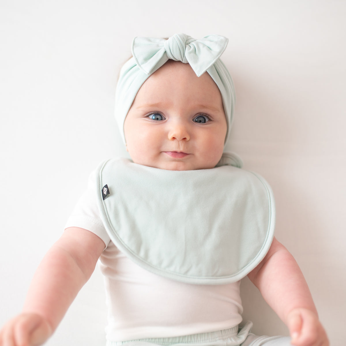 Infant model wearing the Knotted Bow Headband in Dew with matching Cushy bib and a Cloud Bodysuit and