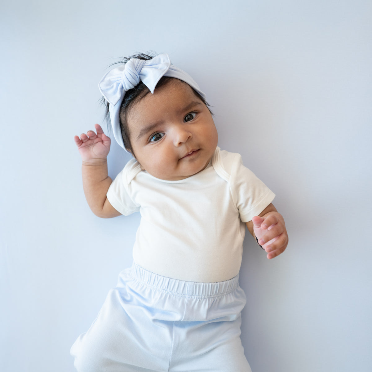 Infant girl wearing the Knotted Bow Headband in Mist paired with matching pants and a Cloud Bodysuit