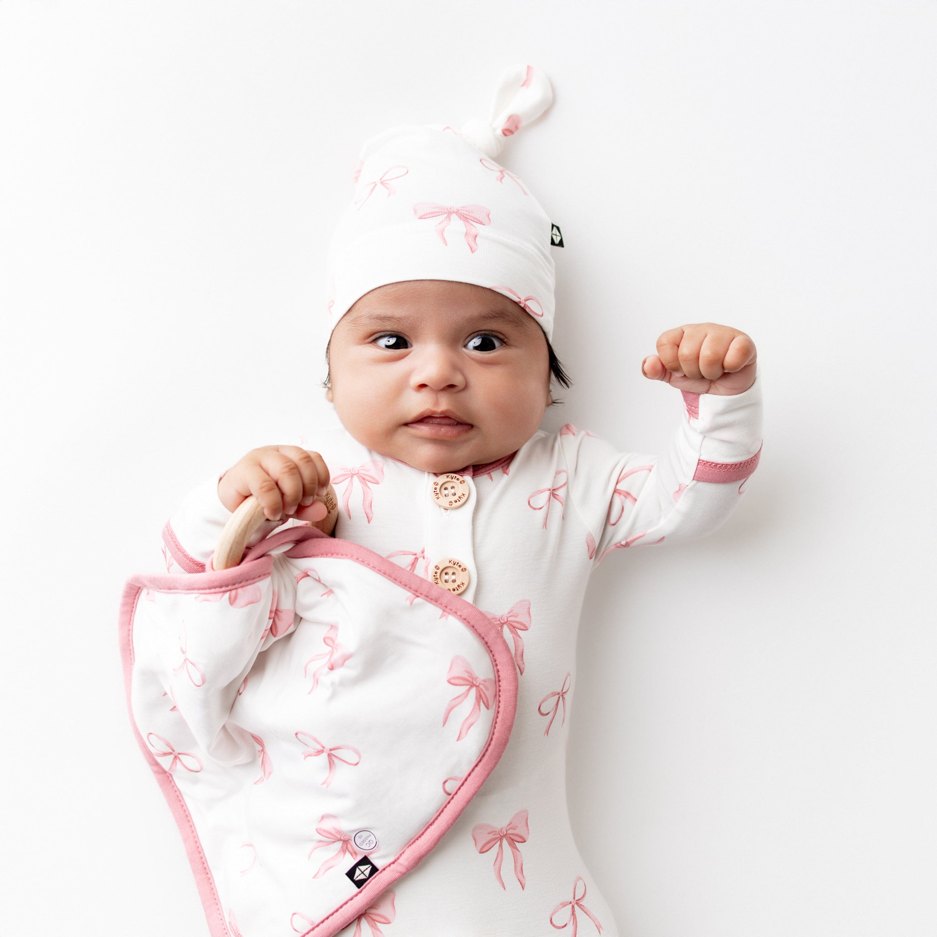 Infant laying on a white surface holding the Lovey in Bow with Removable Wooden Teething Ring wearing a matching knotted gown with hat set