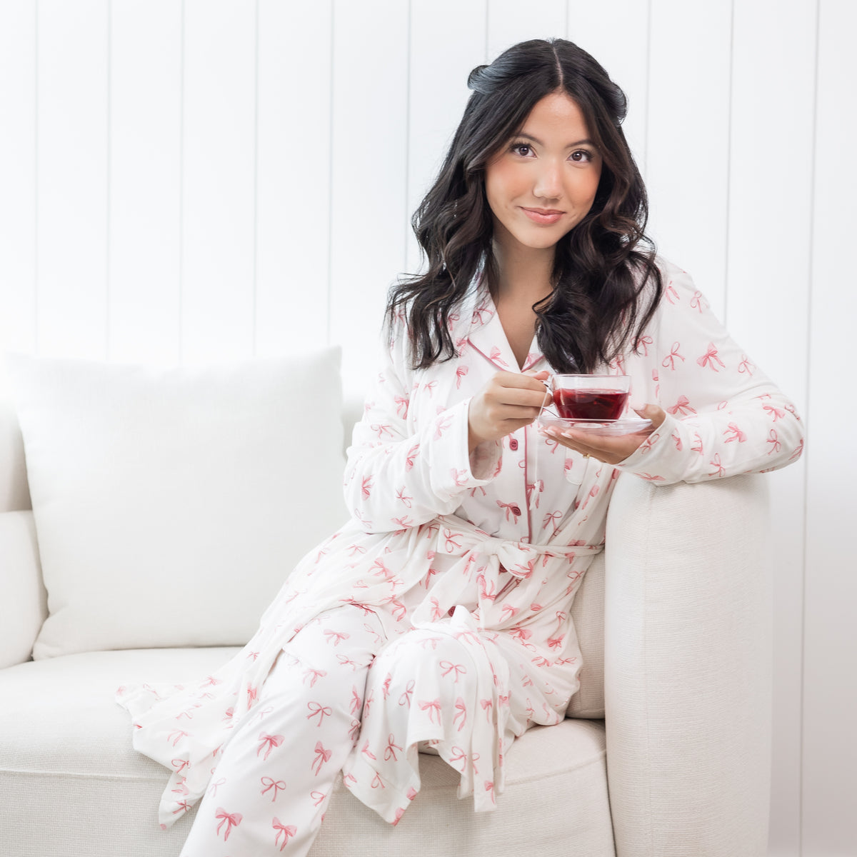 Woman wearing white long sleeve button up pajamas and robe with a pink bow print holding a cup of tea