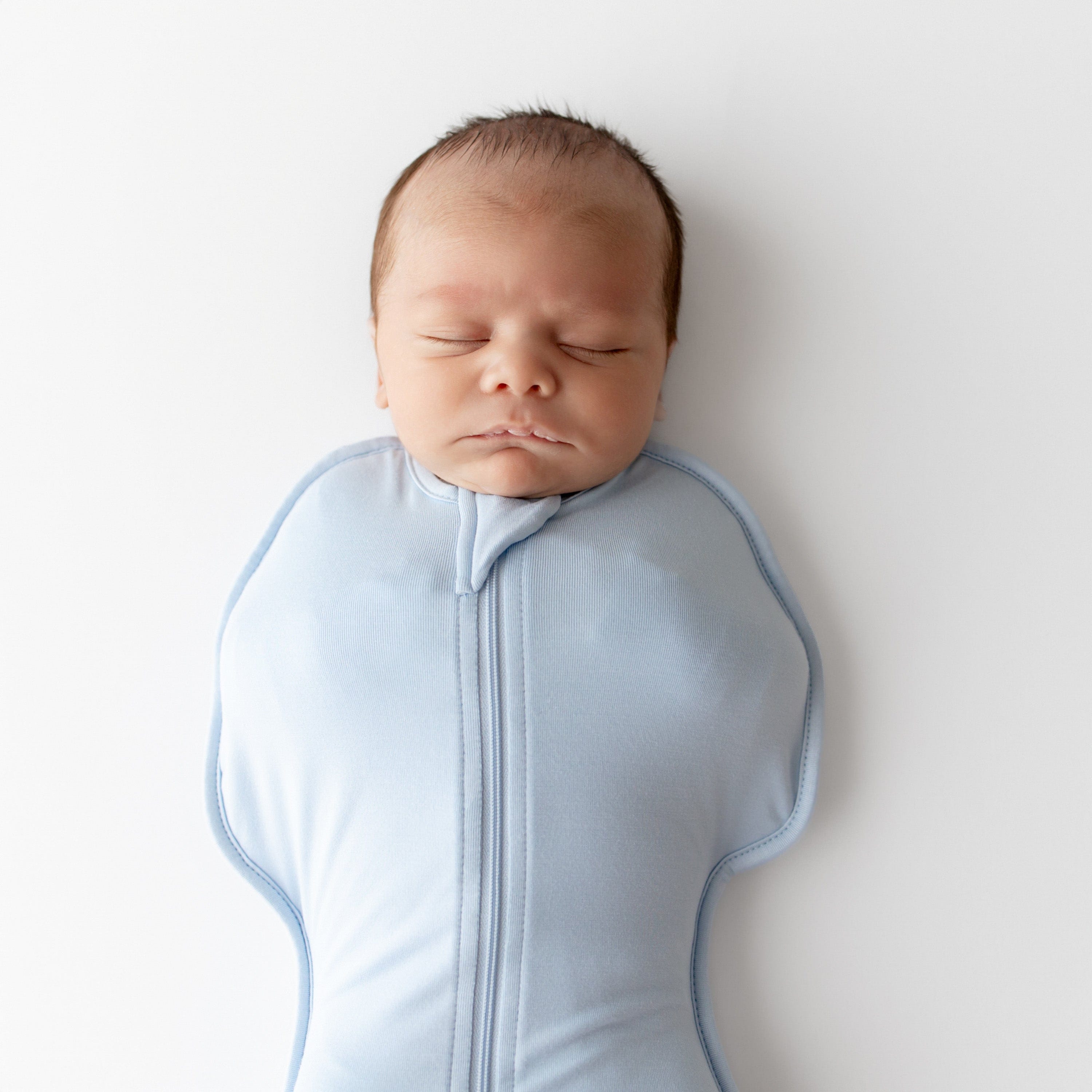 Newborn baby wearing a light blue swaddle bag with zippers on a white background