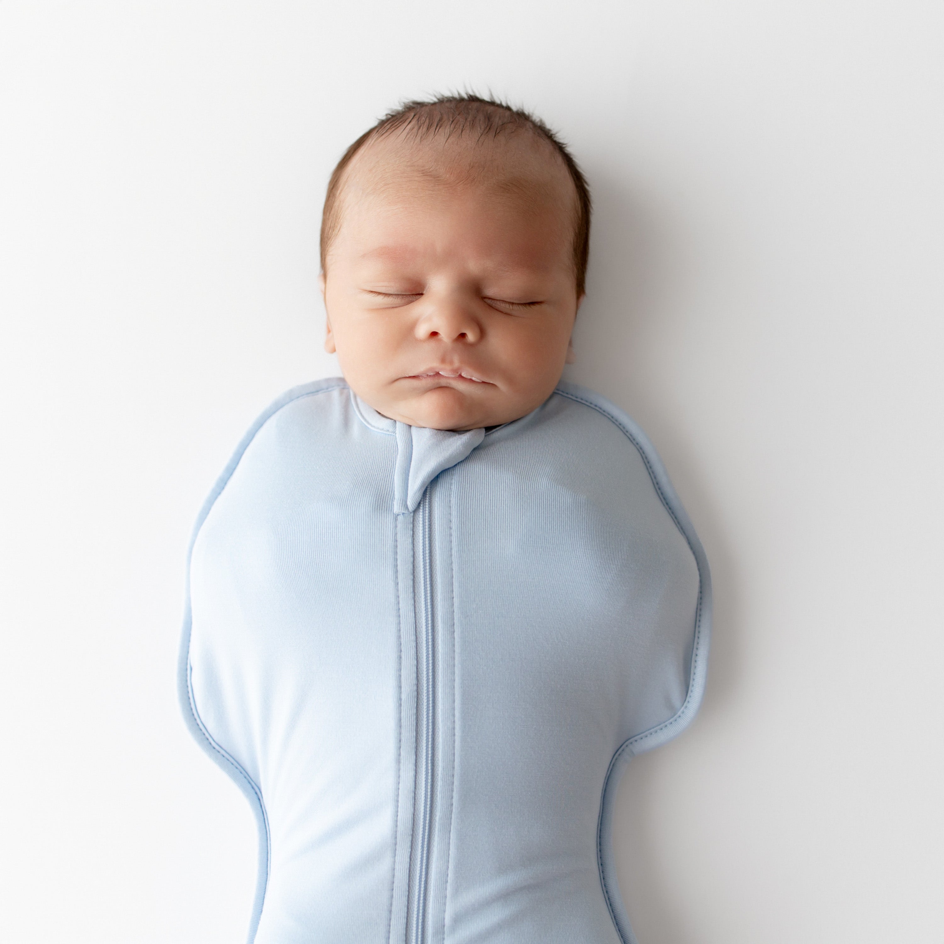 Newborn baby wearing a light blue swaddle bag with zippers on a white background