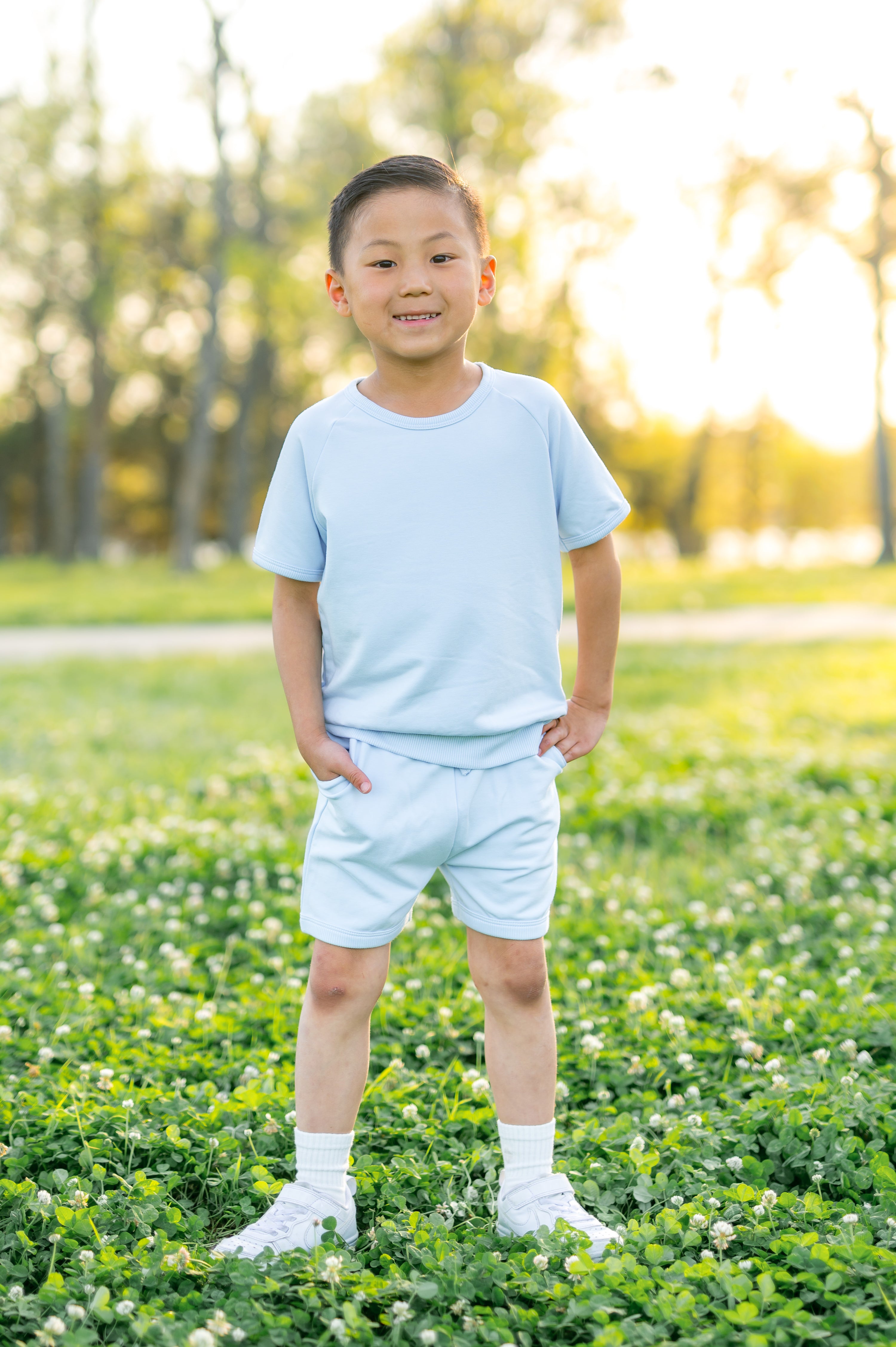 Child in light blue shirt and shorts standing in a grassy field with trees in the background