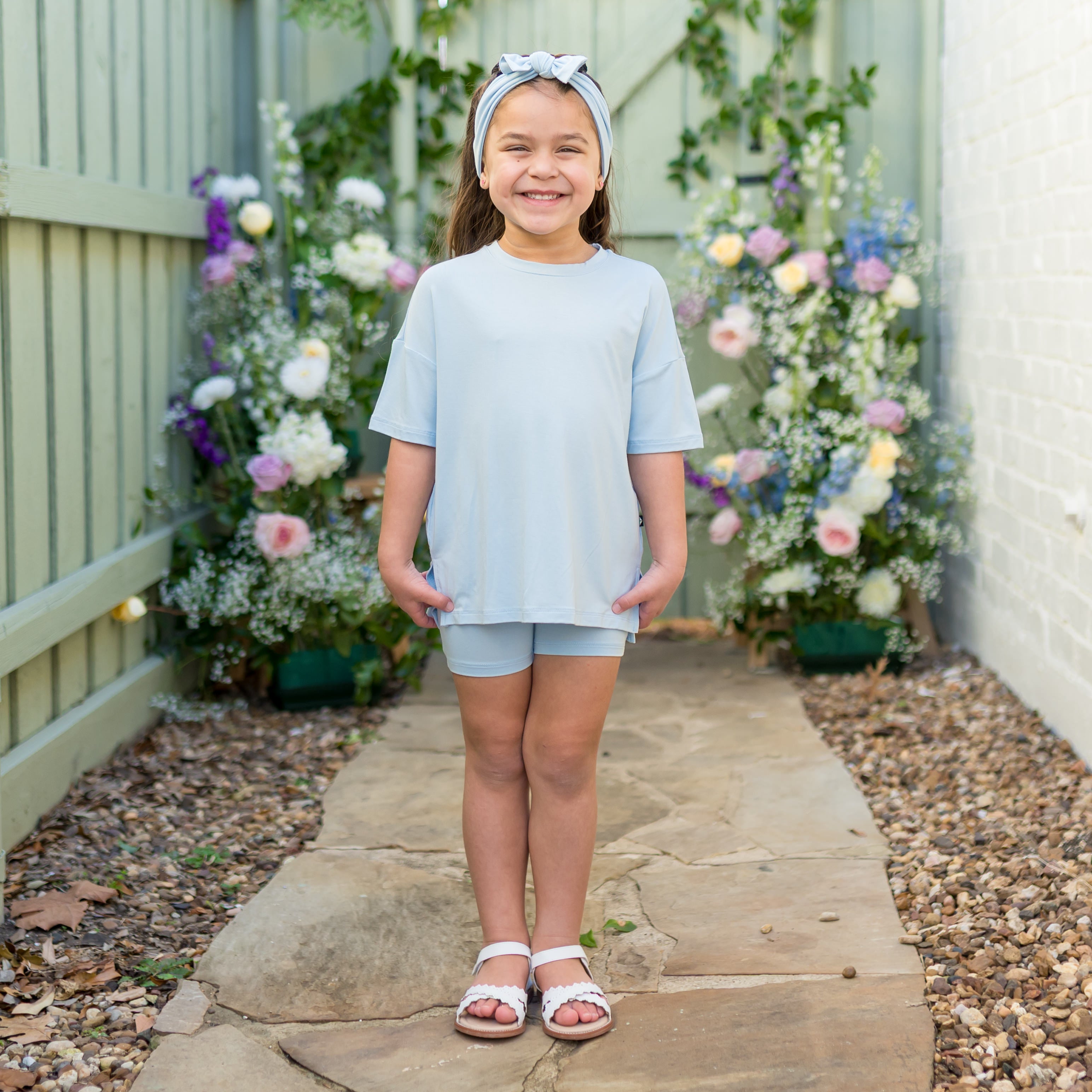 Young girl wearing a light blue biker set standing on a stone path with flowers in the background