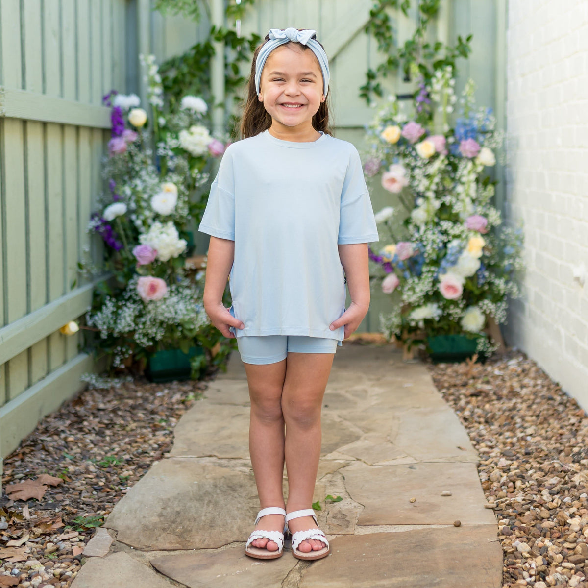 Young girl wearing a light blue biker set standing on a stone path with flowers in the background