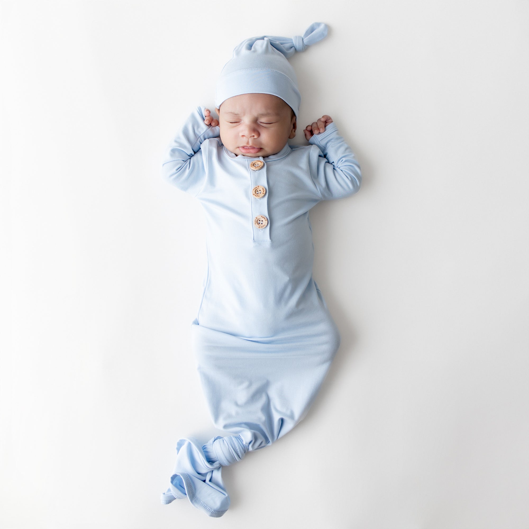 Sleeping newborn wearing the Knotted Gown with Hat Set in Breeze laying on a white surface