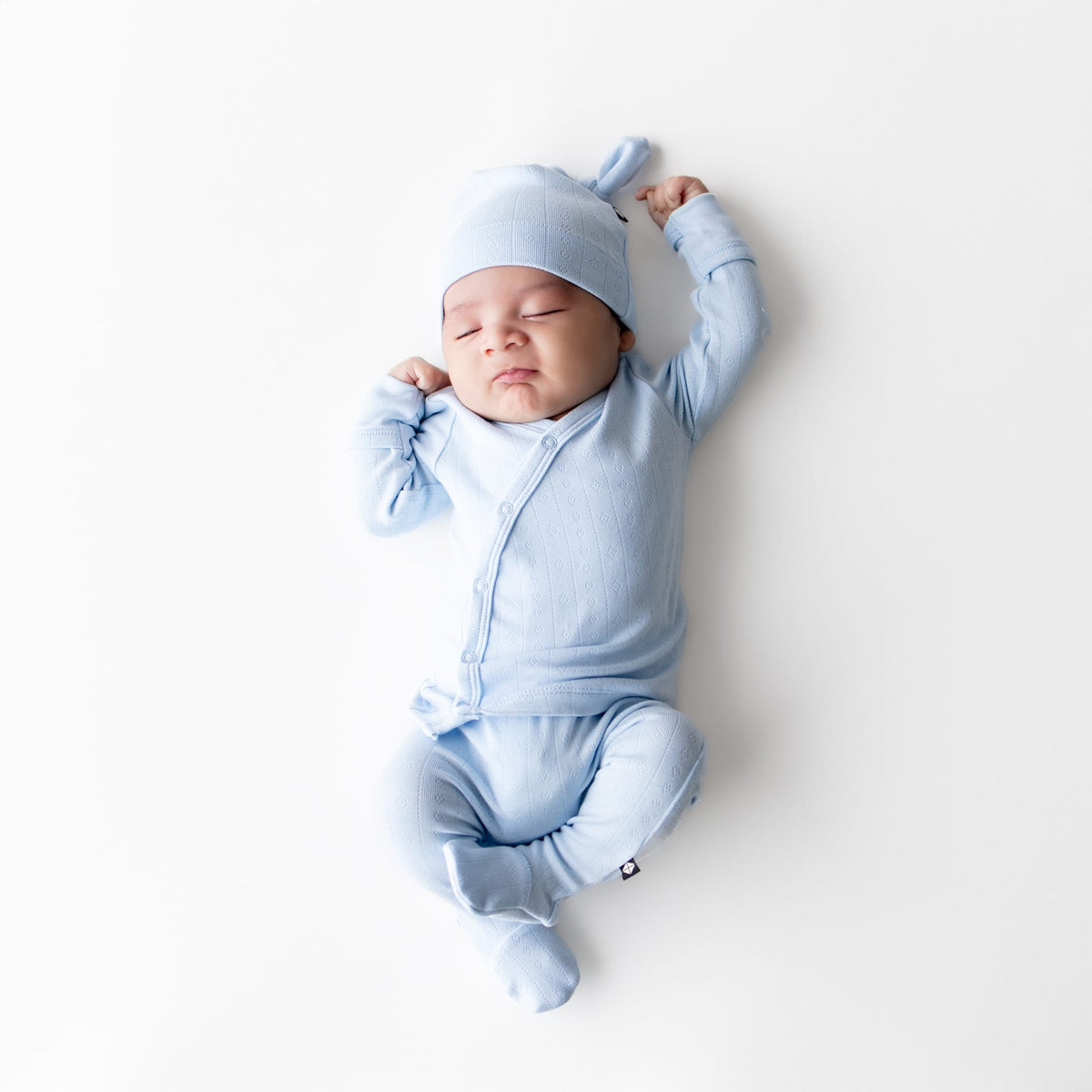 Sleeping newborn laying on a white surface wearing the Pointelle Take Me Home Set with Cap in Breeze
