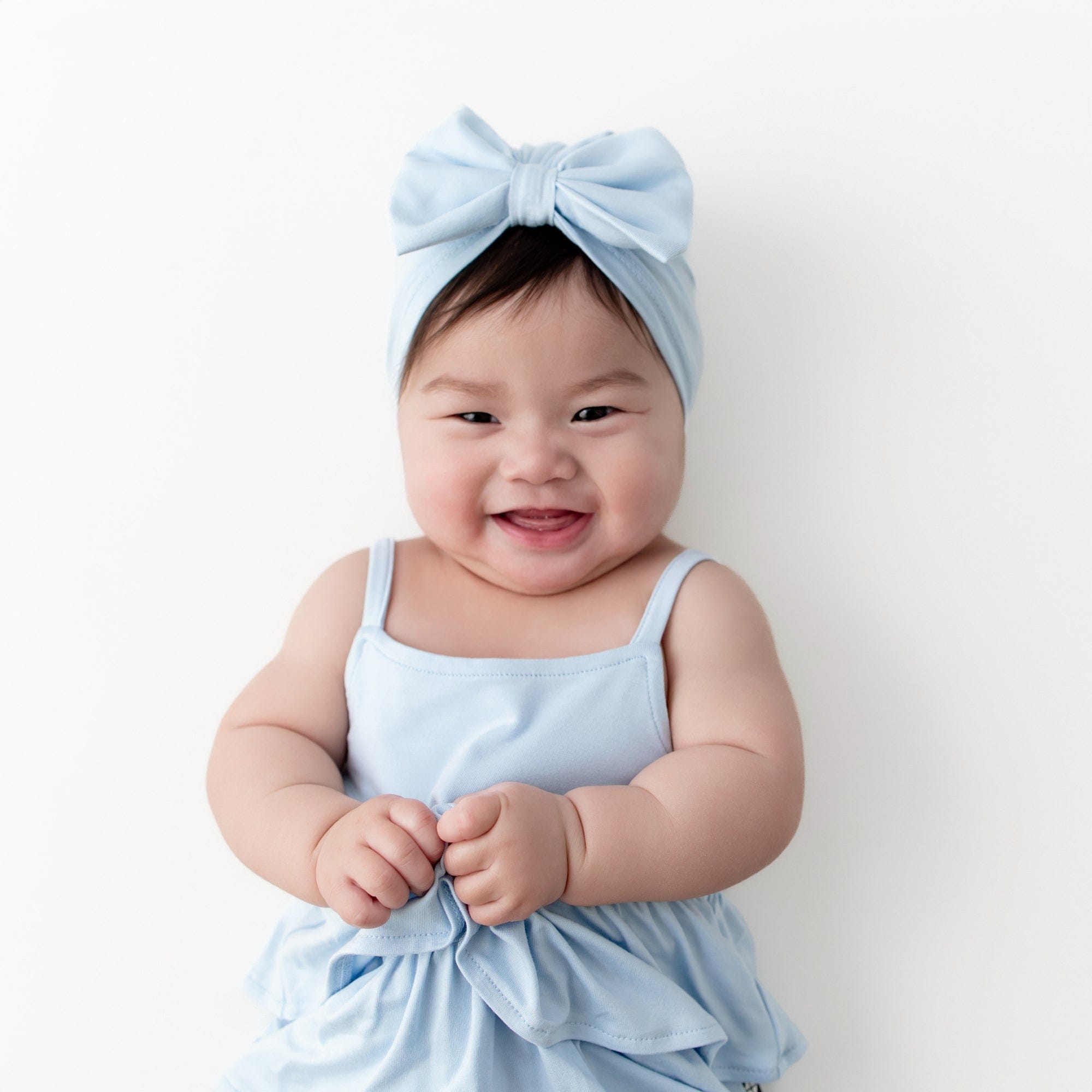 Smiling infant wearing the Bow Headwrap in Breeze with matching spaghetti strap ruffle romper