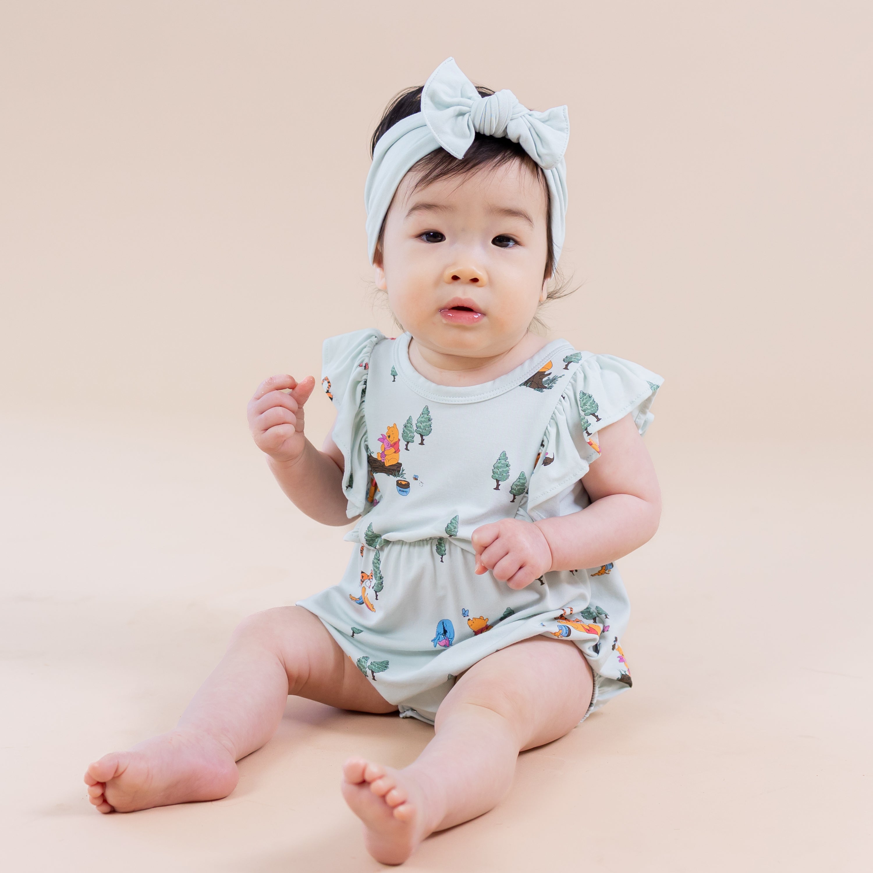 Young toddler sitting on the floor wearing the Bubble Romper in Winnie the Pooh Hundred Acre Wood paired with a light green bow