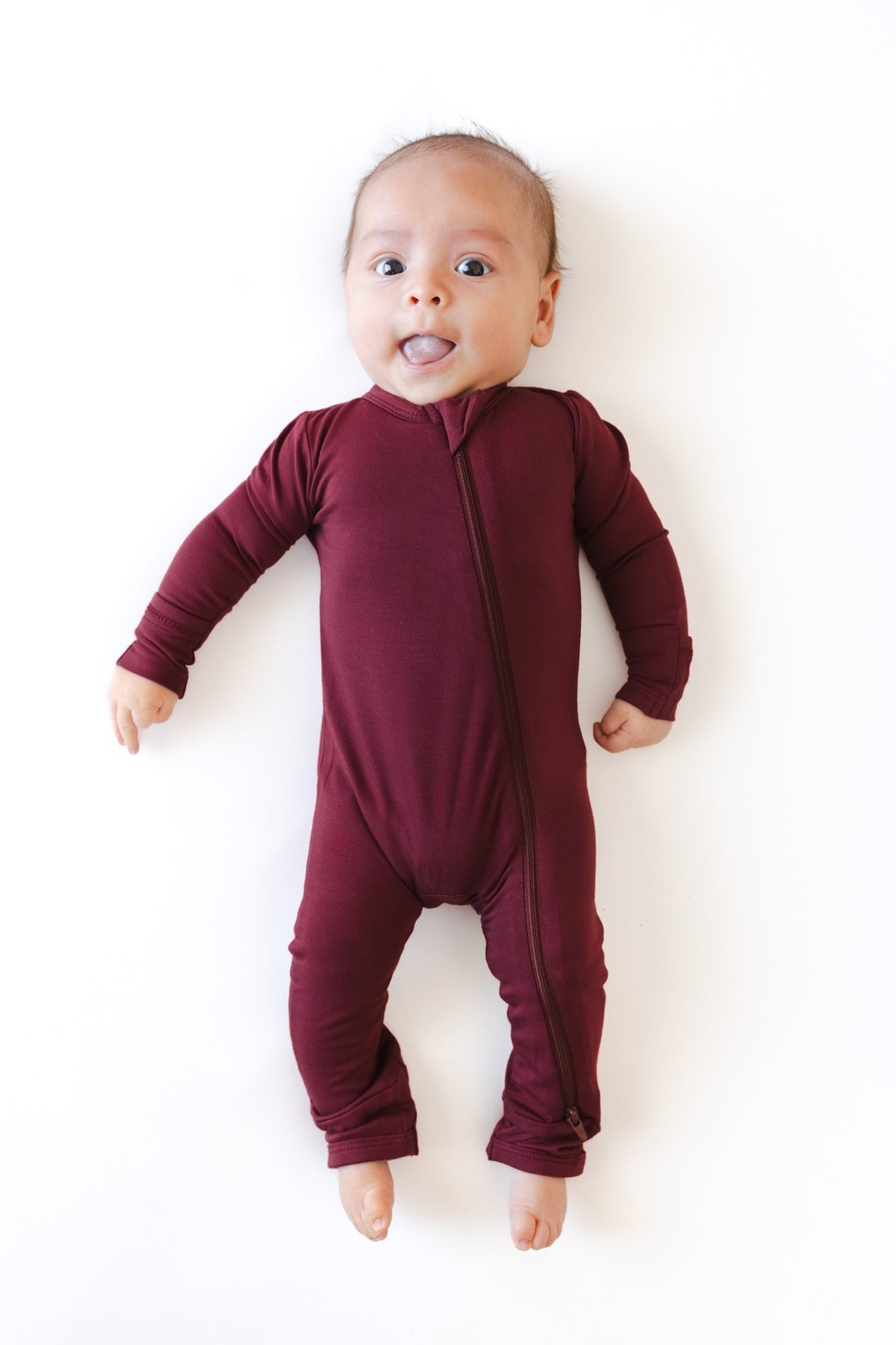 A baby lying on their back wearing a long-sleeve, footless zip-up romper in a deep burgundy color, set against a plain white background.