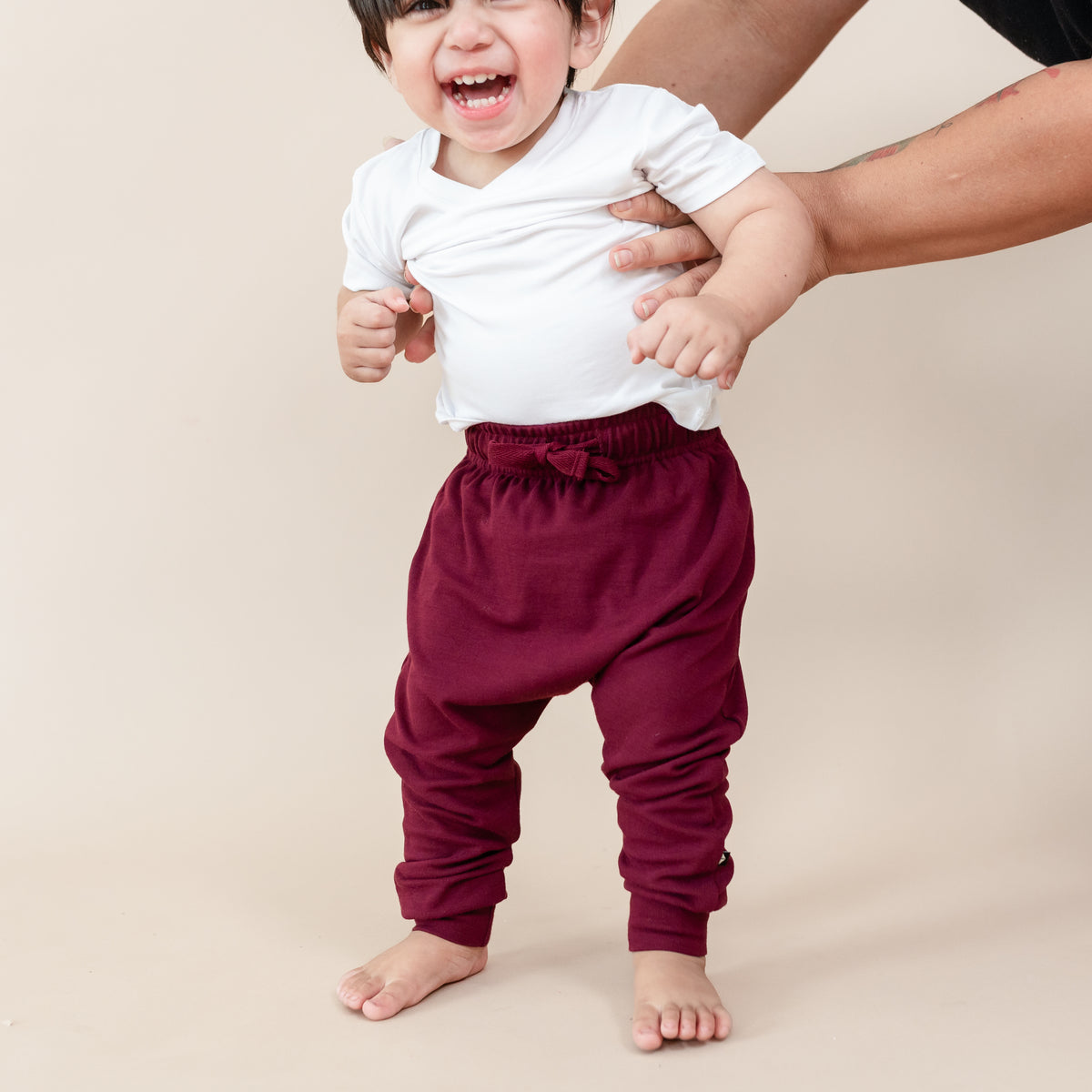 Toddler being held upright wearing the Bamboo Jersey Harem Pants in Burgundy paired with a Snow toddler V-neck in front of a light neutral background