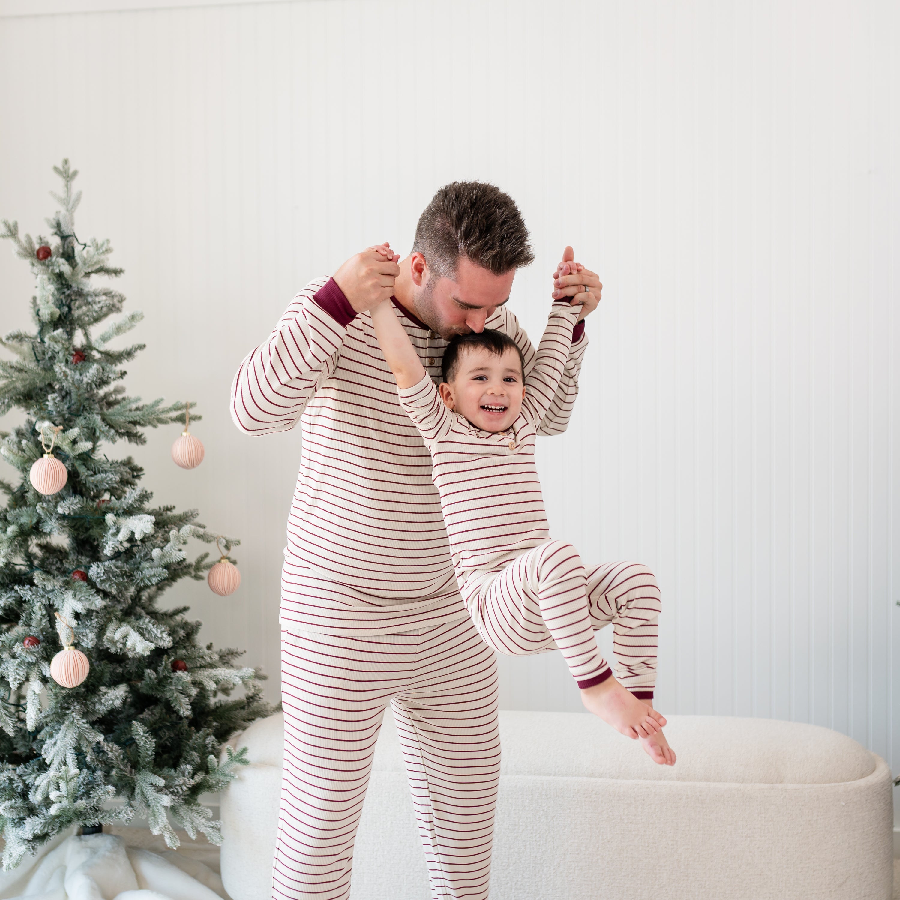 Father wearing the Men's Ribbed Henley Set in Burgundy Stripe holding his son up in the air by the hands kissing him on the head who is in matching ribbed toddler set
