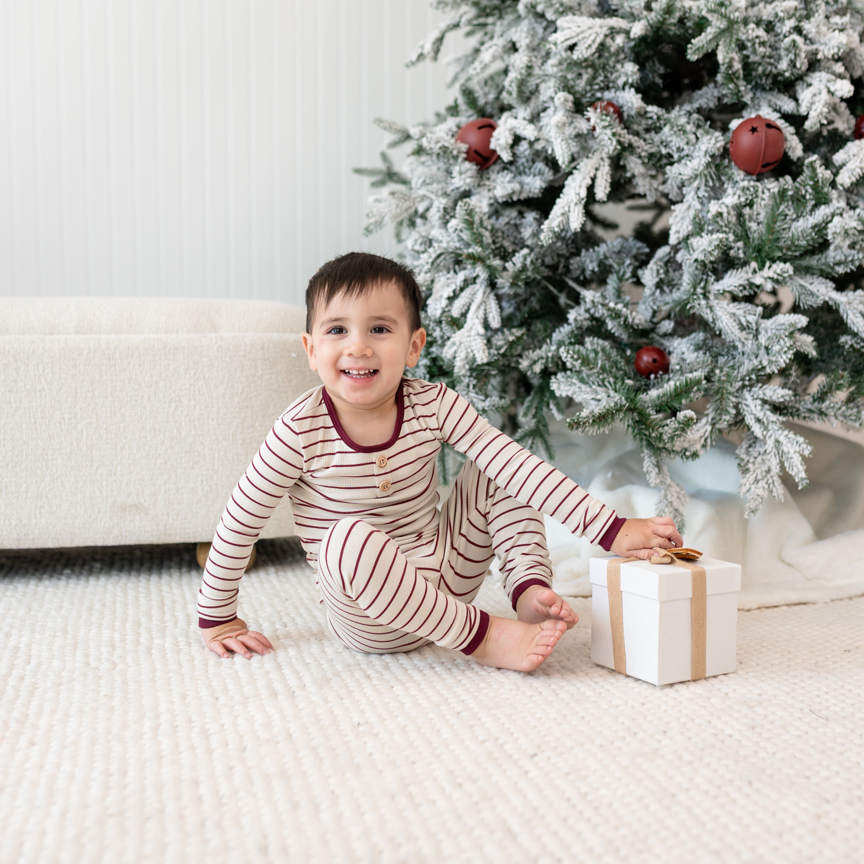 Smiling young boy sitting on a cream carpet with a white box wrapped in a gold bow in front of a decorated christmas tree and cream ottoman wearing the Ribbed Henley Set in Burgundy Stripe