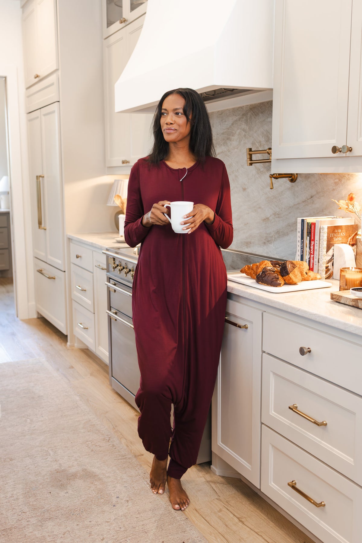 A woman wearing a burgundy sleep sack stands barefoot in a bright white kitchen, holding a white mug.