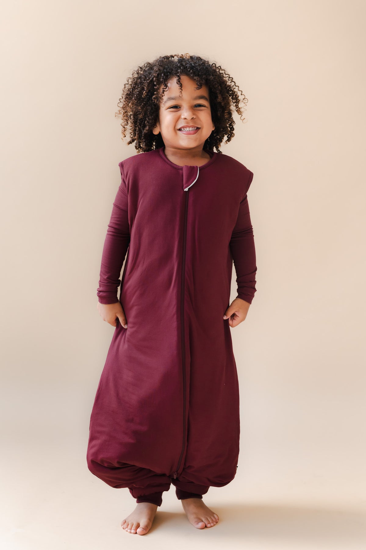 A smiling toddler stands barefoot against a plain beige studio background, wearing a full-length zip-front sleep sack and pajama in deep burgundy