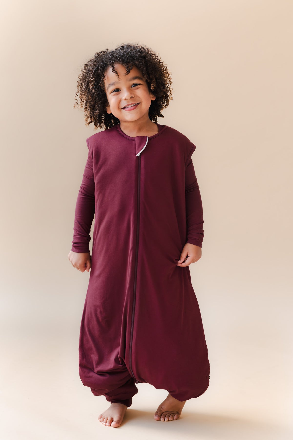 A smiling young child, wearing a full-length zip-front sleep sack and pajama in deep burgundy. The garment has a loose harem-style leg silhouette, and a small light zipper.