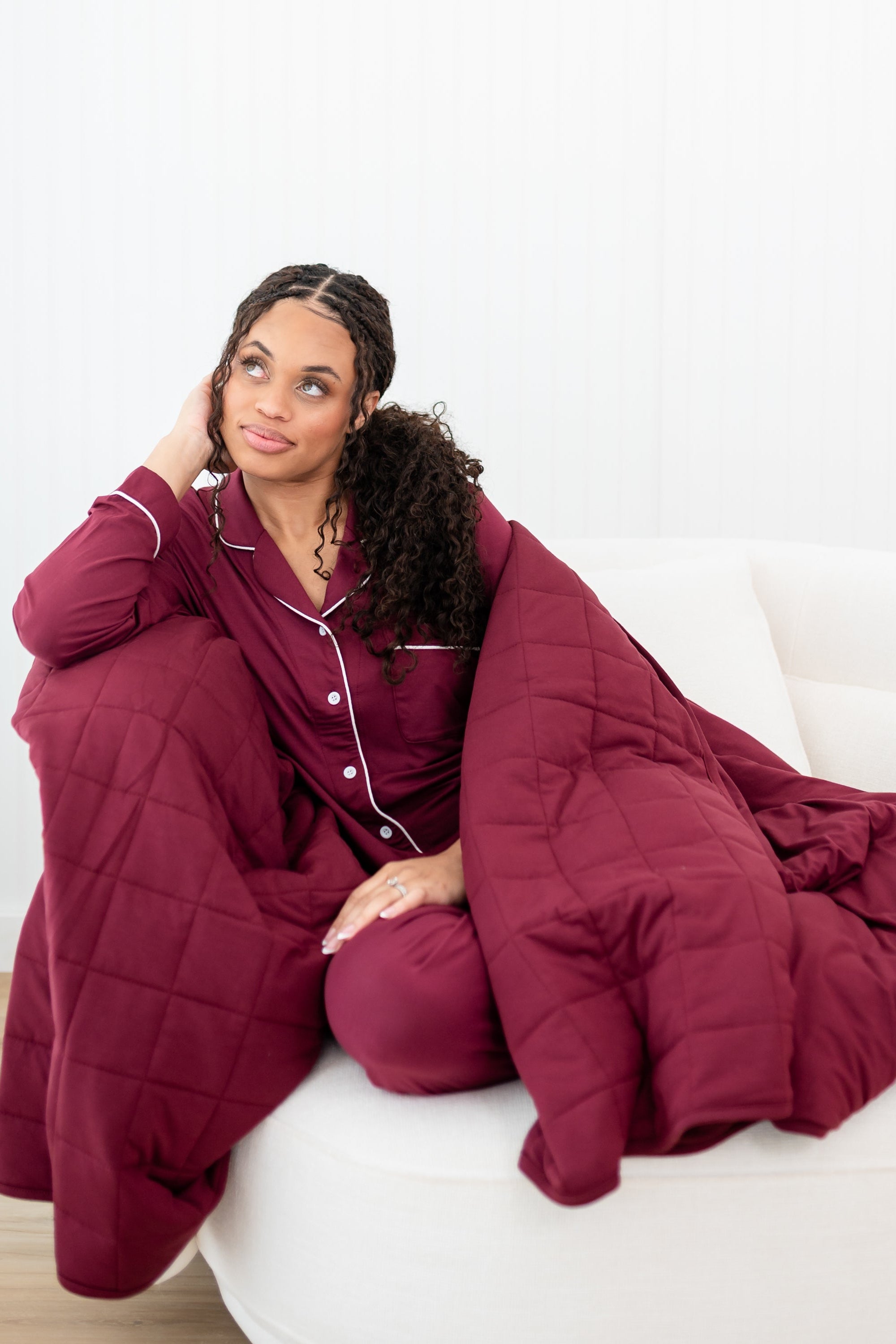Female sitting on a couch wrapped in the Adult Quilted Blanket in Burgundy 2.5 wearing matching long sleeve women's pajama set