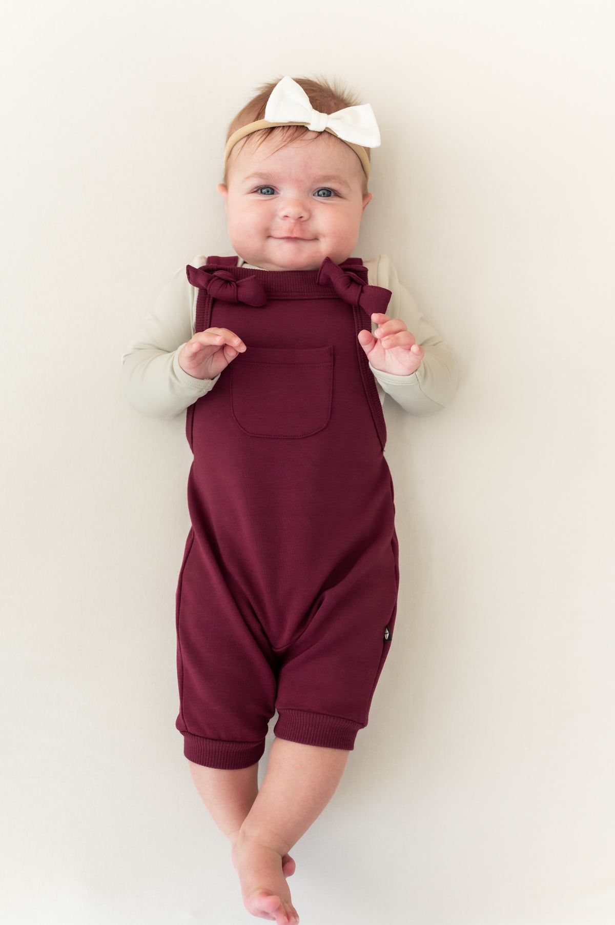 Baby wearing Overall in Burgundy color with white bow headband lying on a beige background