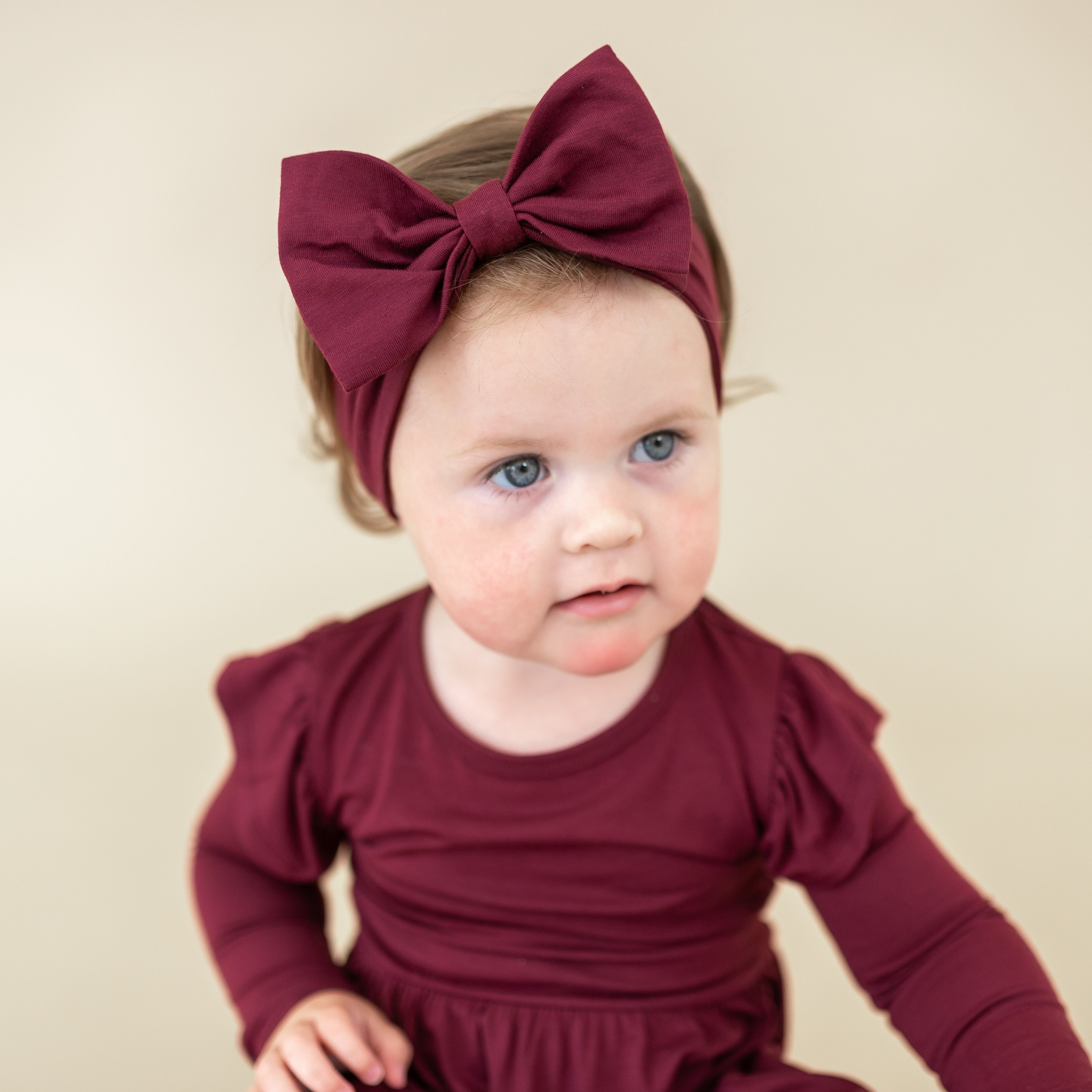 Infant looking off to the side wearing the Bow Headband in Burgundy with matching long sleeve twirl bodysuit dress