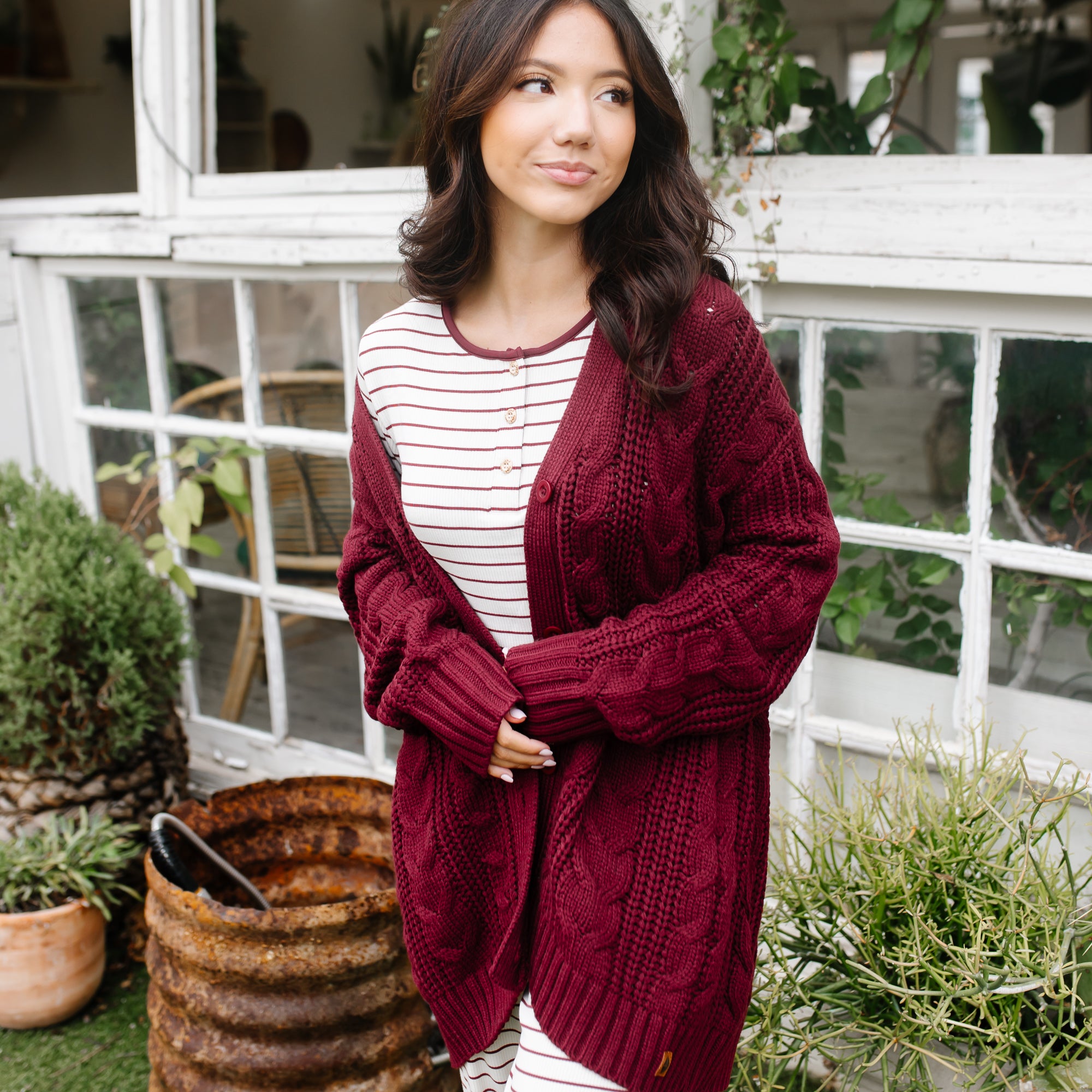 Female modeling the Cable Knit Adult Cardigan in Burgundy wearing the Women's Ribbed Burgundy Stripe Henley Set underneath standing in front of white trimmed windows and potted plants