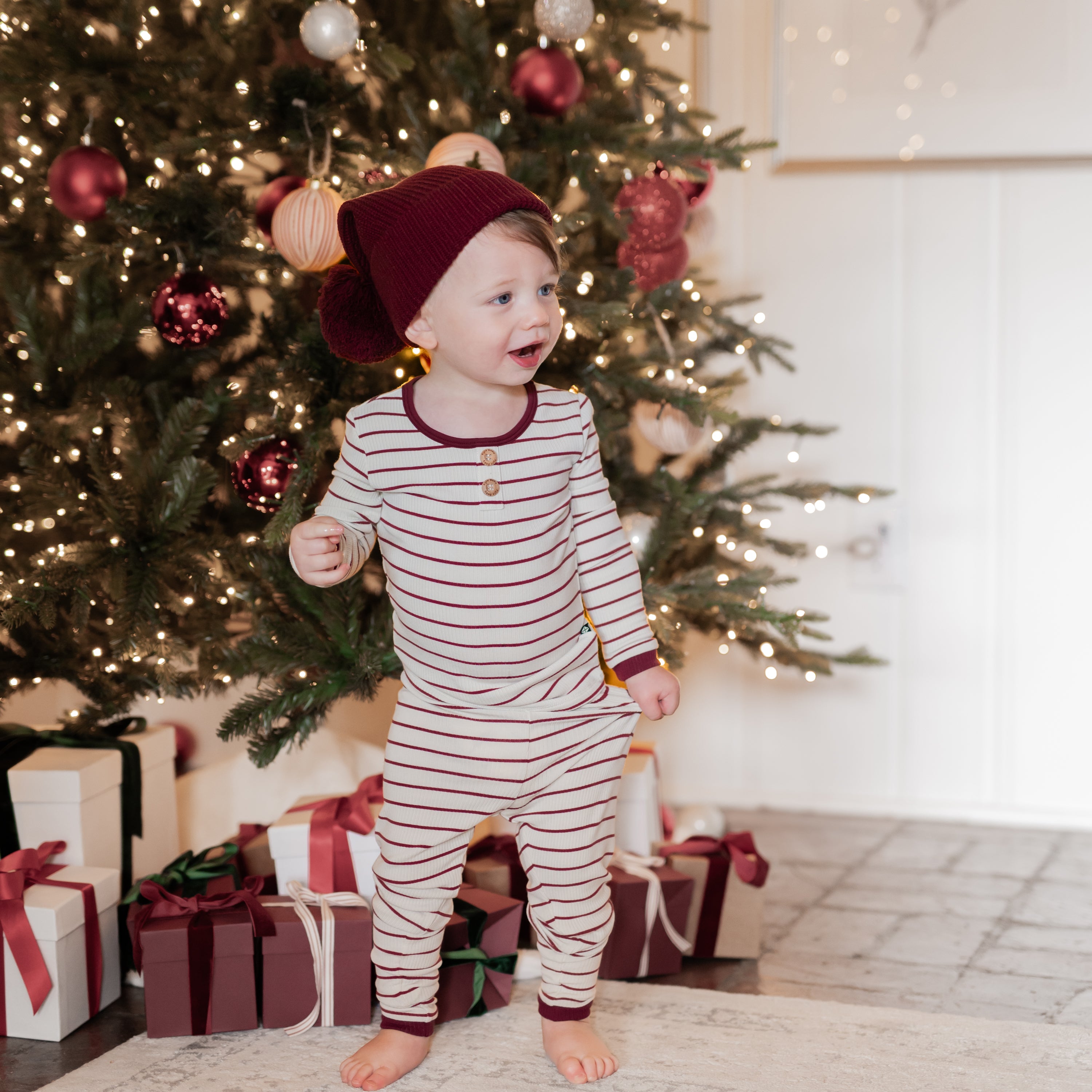 Young toddler standing in front of a decorated christmas trees with gifts underneath wearing the Ribbed Henley Set in Burgundy Stripe and burgundy chunky knit pom beanie