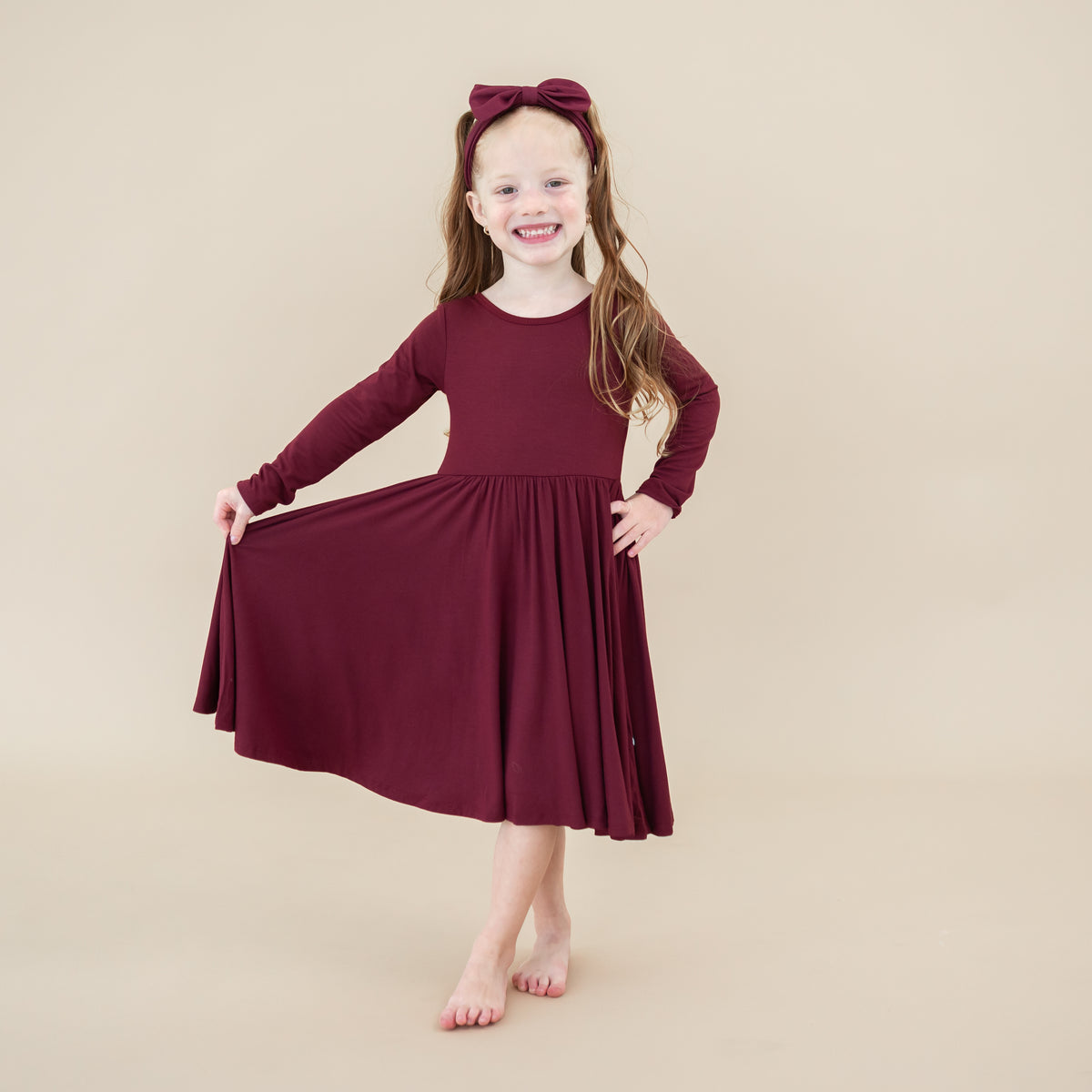 Toddler standing holding one side of the Long Sleeve Twirl Dress in Burgundy out