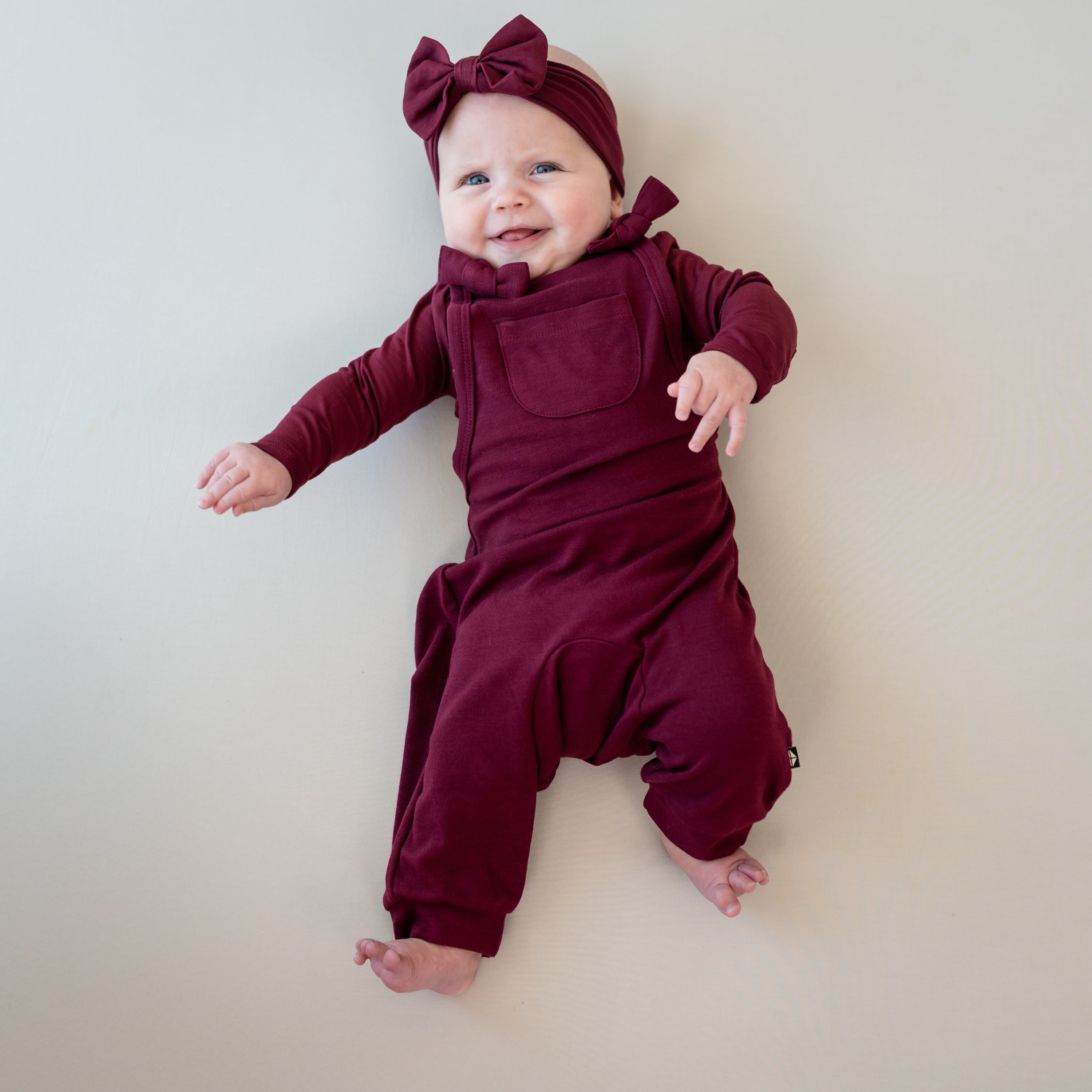 Smiling infant girl laying on a light neutral background with her hands in the air wearing the Bamboo Jersey Overall in Burgundy with matching long sleeve bodysuit and bow headband