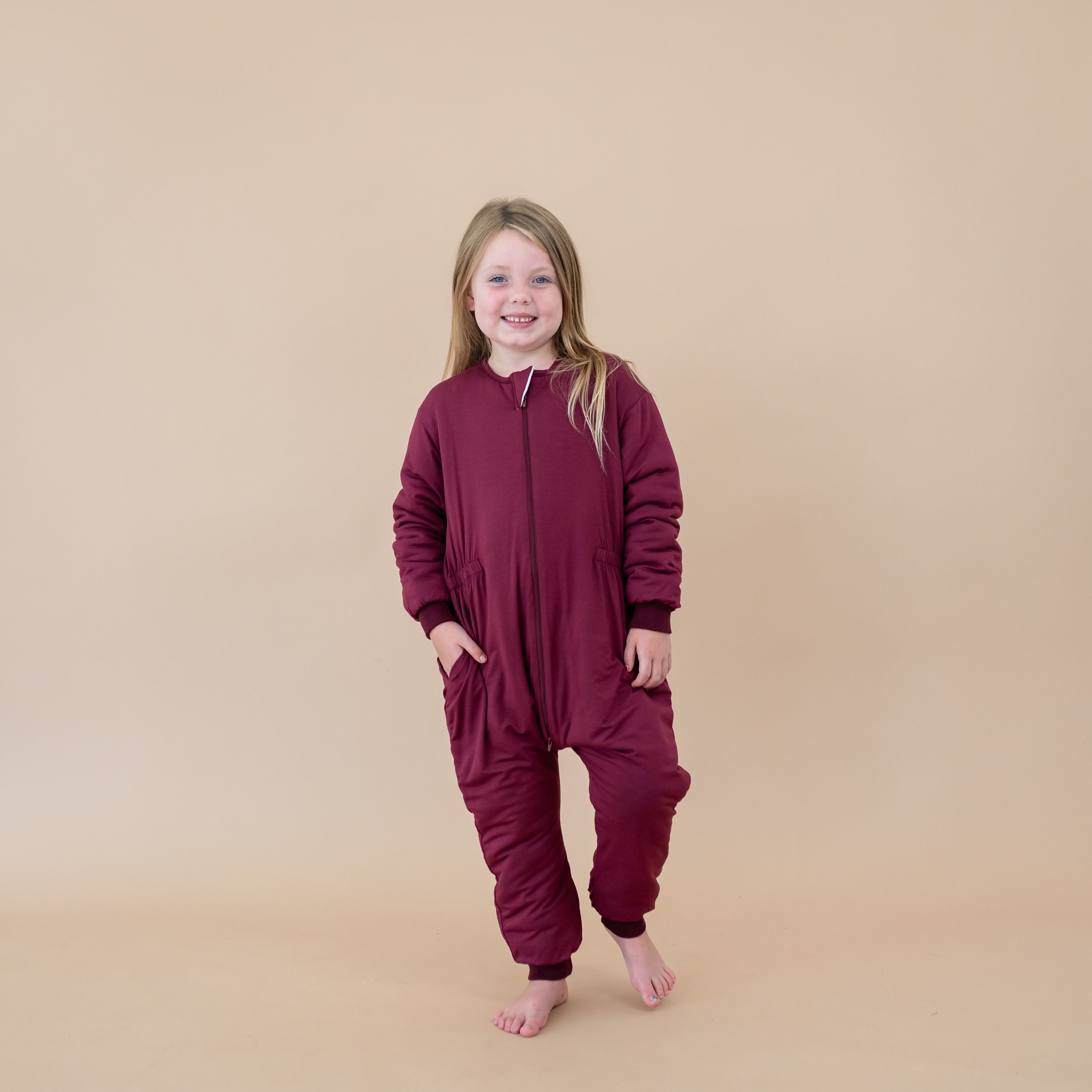 Smiling young girl with one hand in the pocket of the Cozy Playsuit in Burgundy standing in front of a light taupe backdrop