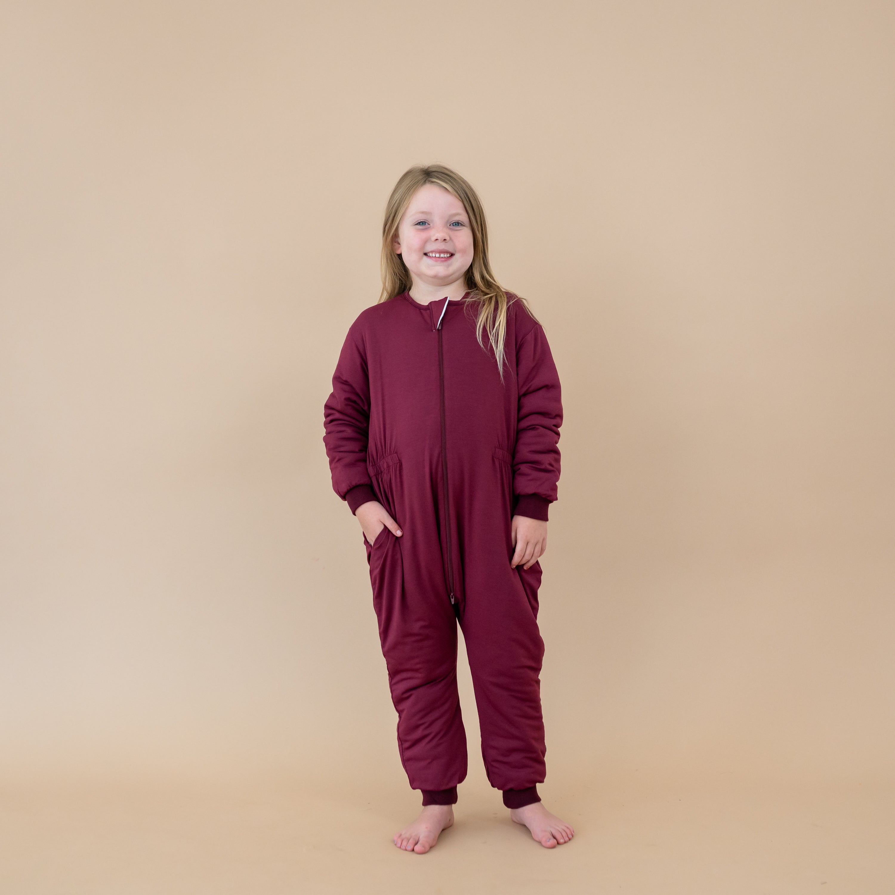 Smiling young girl with on hand in the pocket of the Cozy Playsuit in Burgundy standing in front of a light taupe backdrop