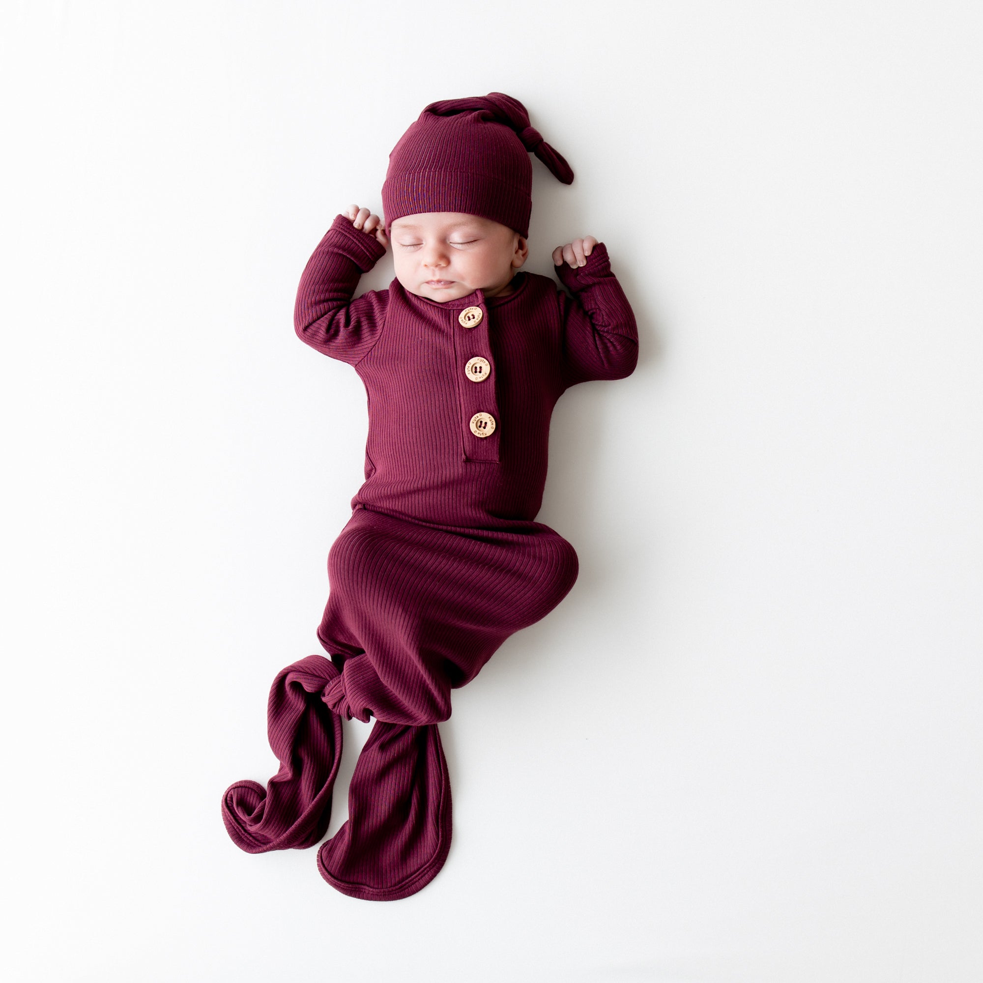 Sleeping infant wearing the Ribbed Knotted Gown with Hat Set in Burgundy laying on a light neutral background