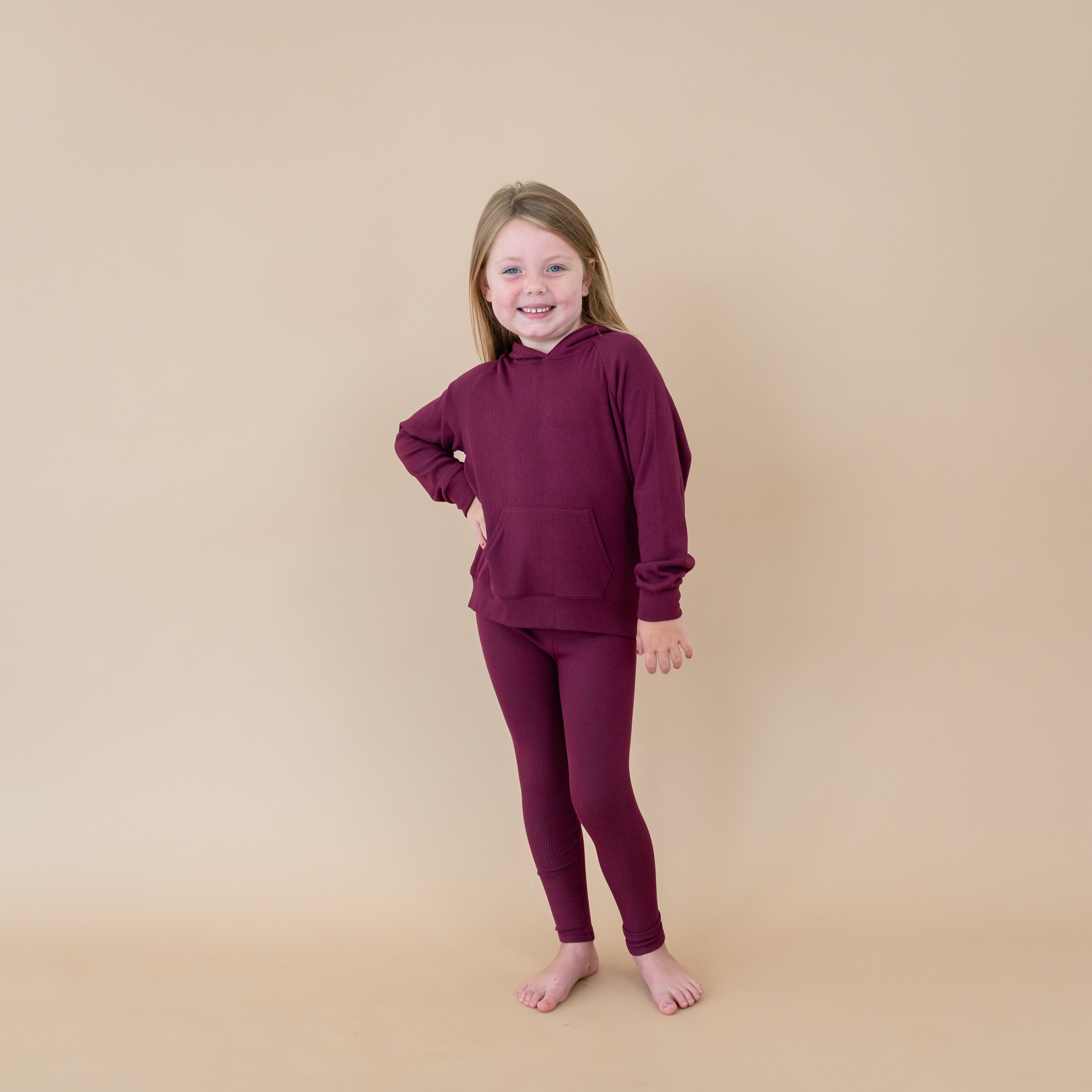 Smiling girl model standing with her hand on her hip wearing the Ribbed Hoodie in Burgundy paired with matching ribbed leggings in front of a taupe backdrop