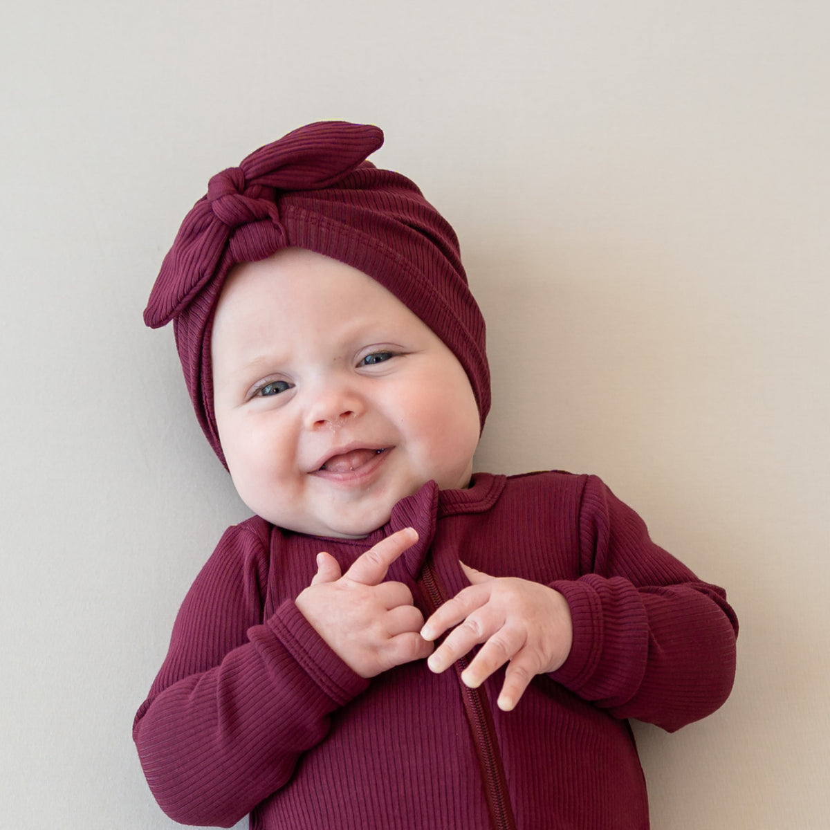 Smiling infant wearing the Ribbed Headwrap in Burgundy with matching ribbed zipper romper