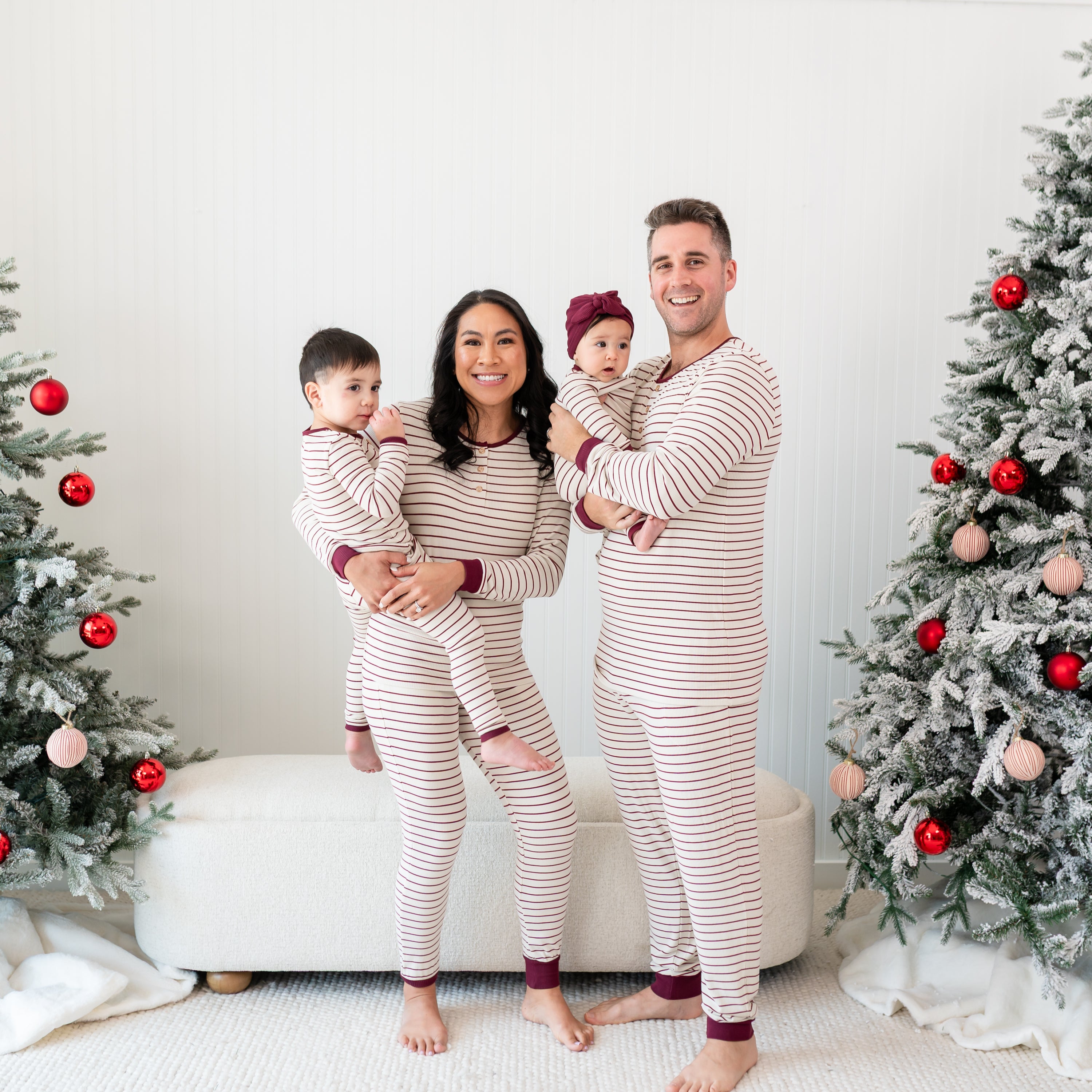 Family of four standing between two decorated Christmas trees wearing matching Ribbed Henley Sets in Burgundy Stripe