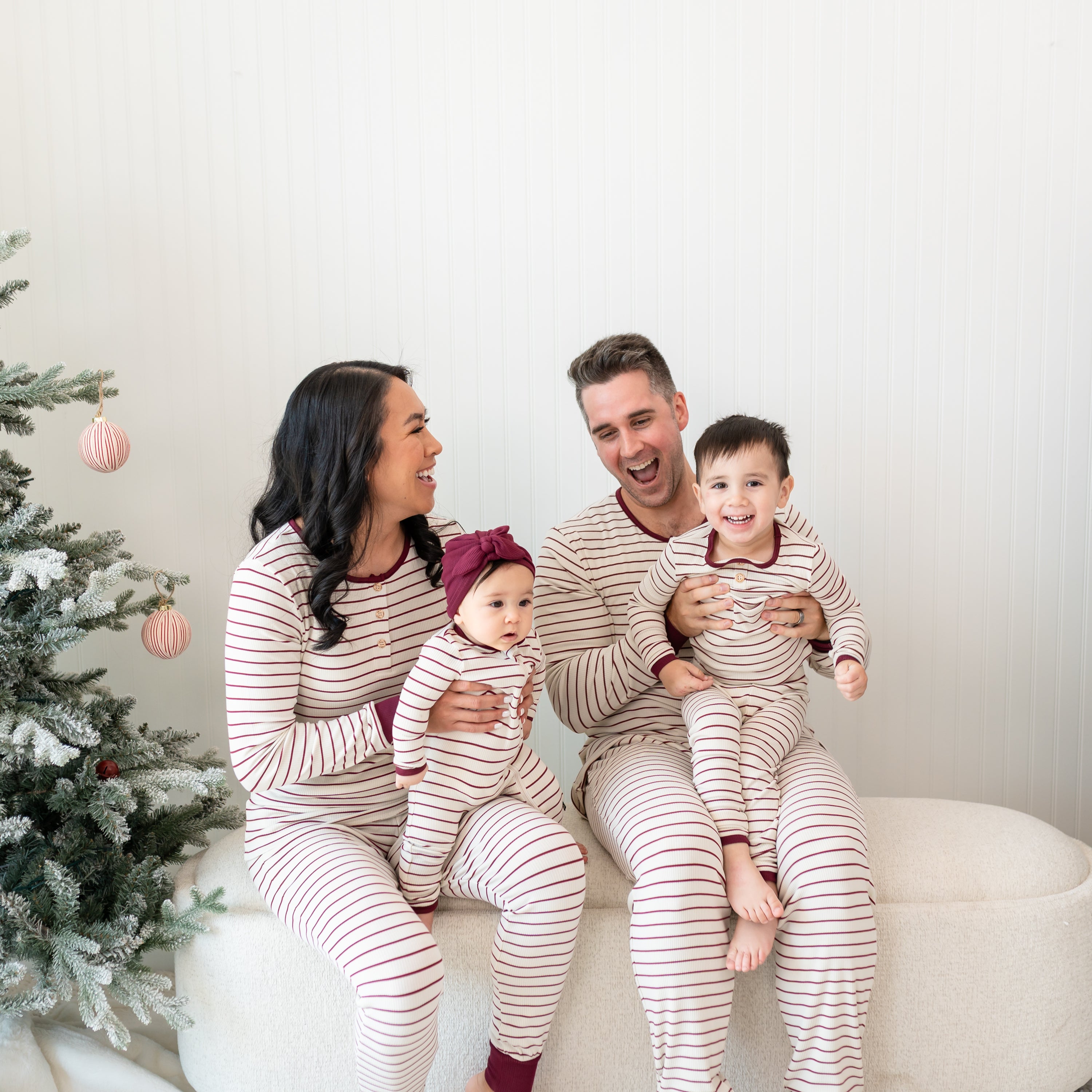 Family of four sitting on a cream ottoman beside a decorate Christmas tree wearing Ribbed Henley Sets in Burgundy Stripe