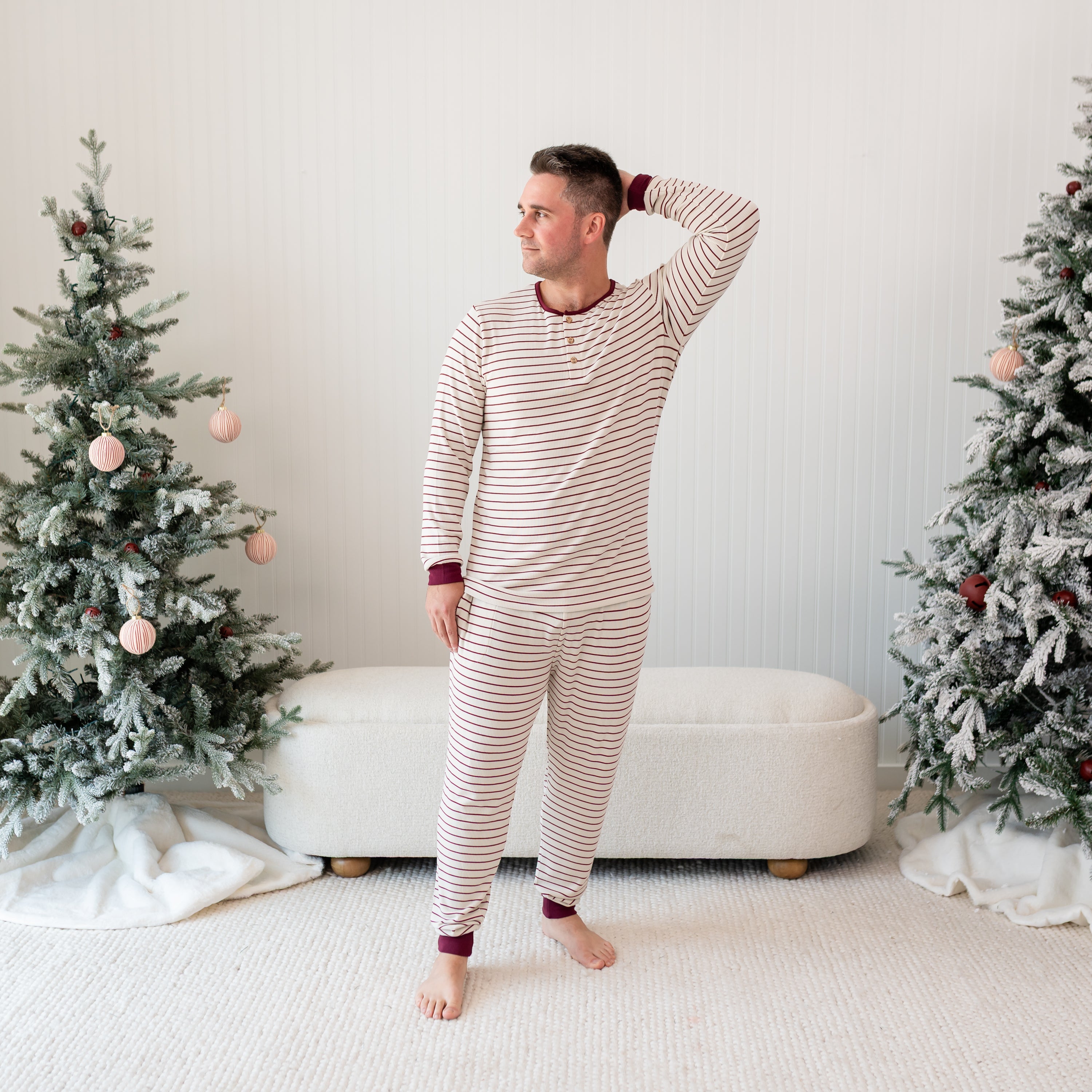 Male model standing in front of a cream ottoman between two christmas trees wearing the Men's Ribbed Henley Set in Burgundy Stripe with one hand behind his head
