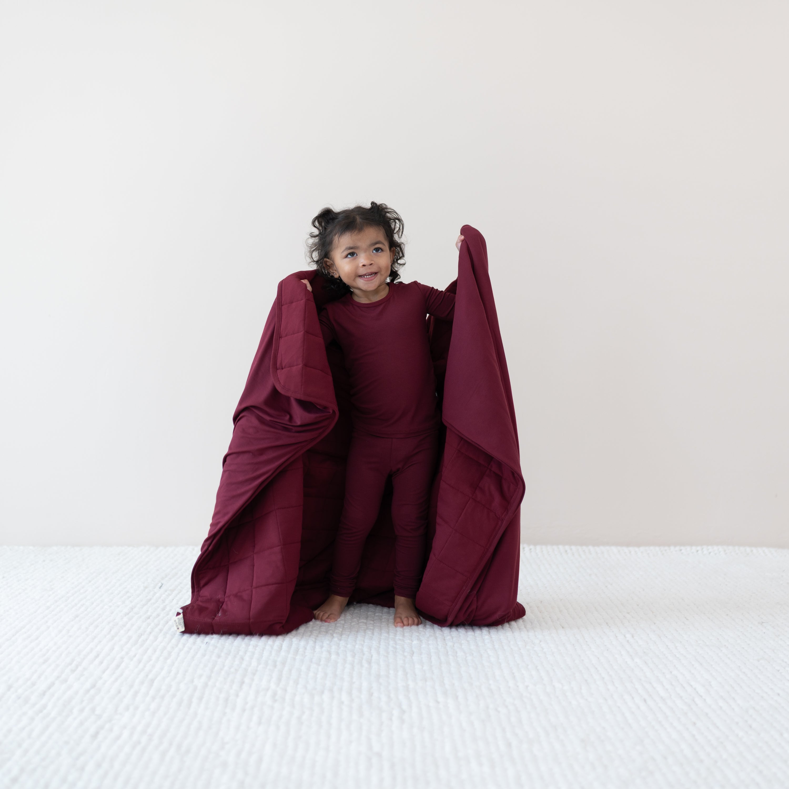 Toddler standing holding the Toddler Blanket in Burgundy 2.5 around her wearing the matching long sleeve toddler pajama set