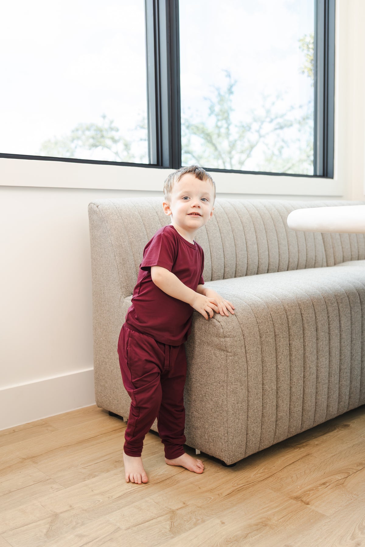 Child wearing Toddler Jogger Pant in Burgundy under Toddler basic tee in burgundy standing against a sofa
