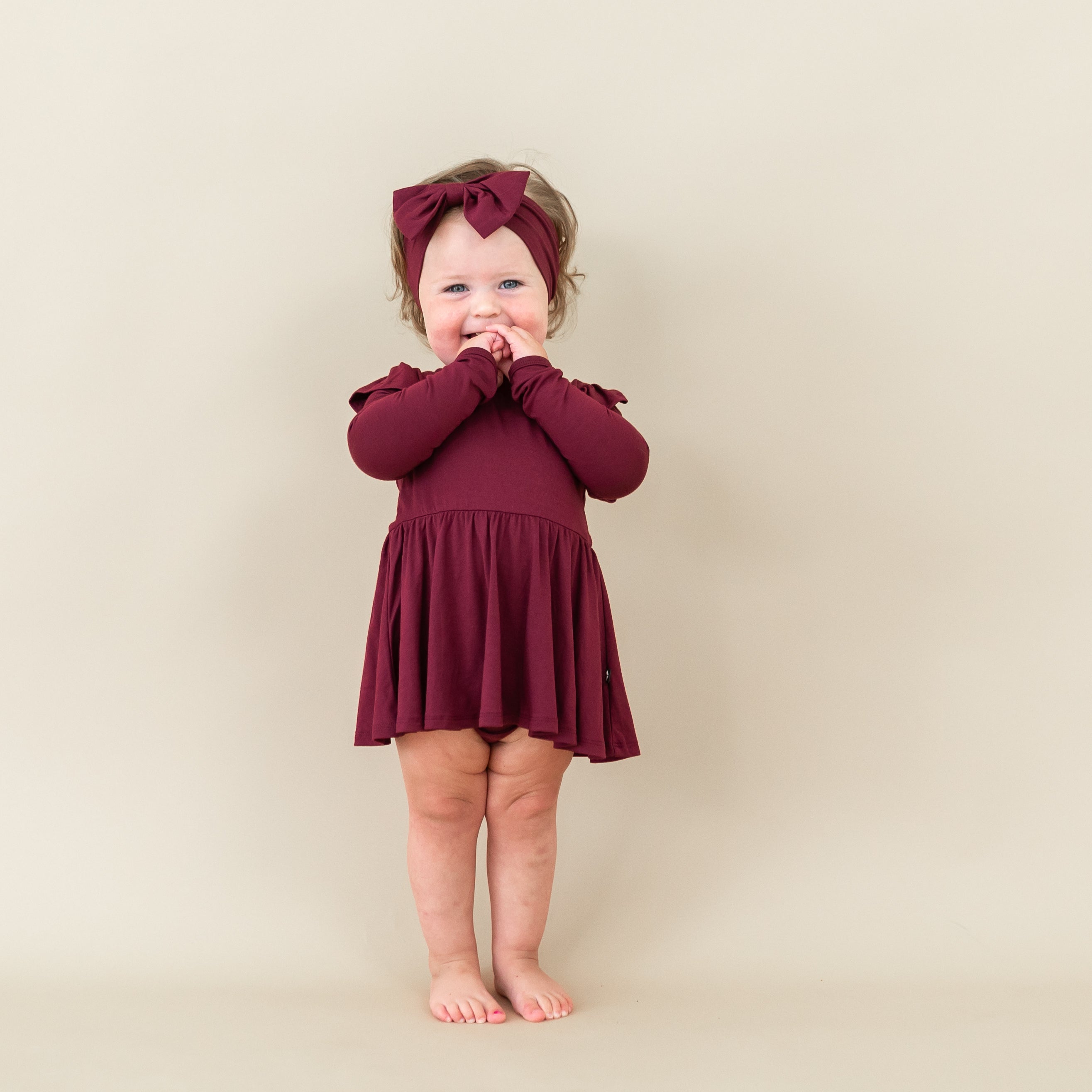 Smiling toddler standing with hands in her mouth wearing the Long Sleeve Twirl Bodysuit Dress in Burgundy and matching bow headband